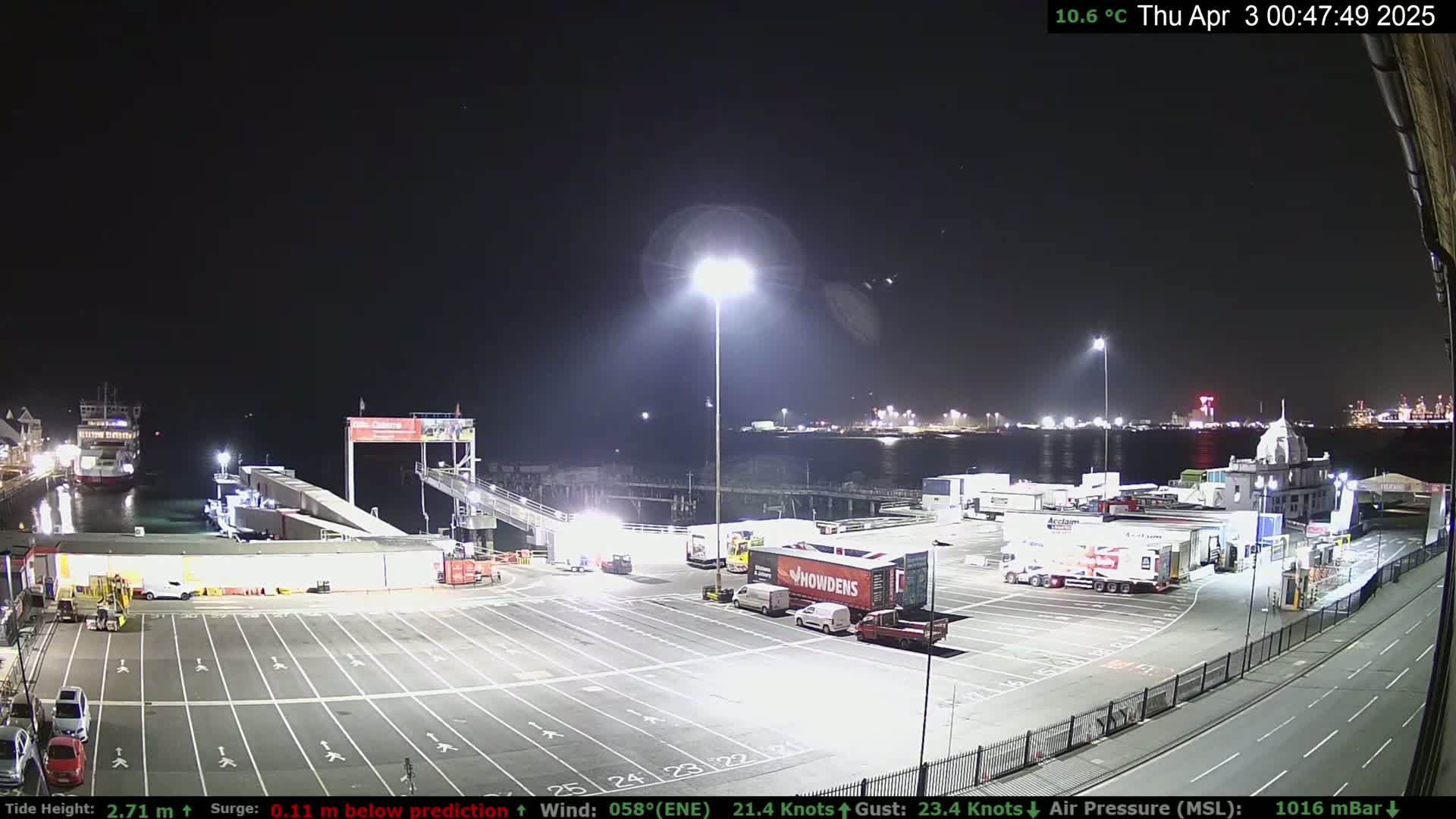 A nighttime view of a ferry terminal with several trucks and cars parked in the lot, a ferry docked at the terminal, and a body of water with lights visible in the distance.