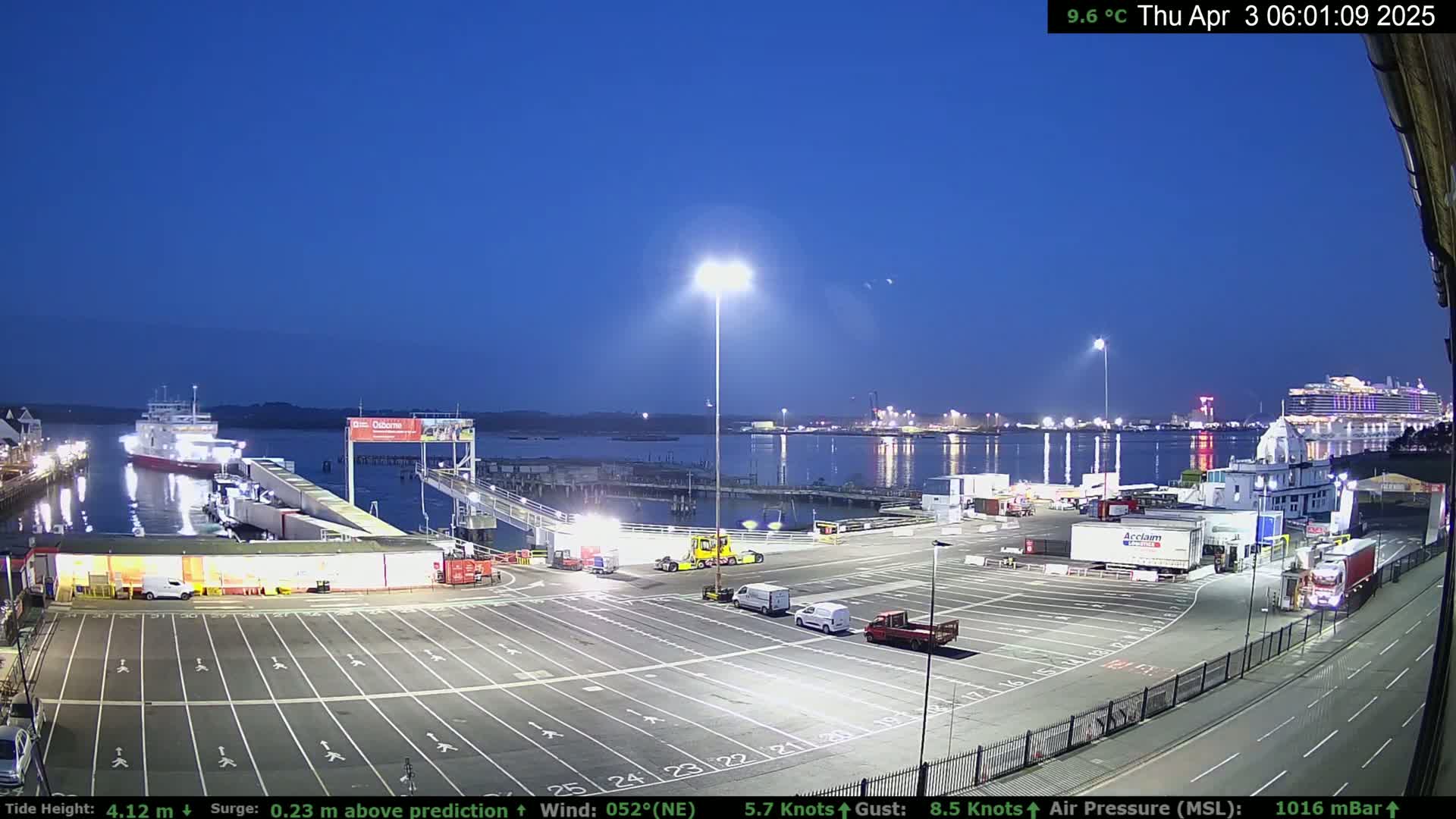 A ferry terminal at twilight, with several vehicles parked and a ferry docked, under a clear, dark-blue sky.