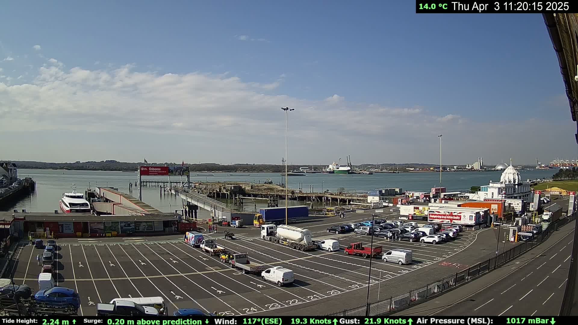 A ferry terminal with numerous cars parked in a lot, a ferry docked at the pier, and calm, sunny weather.