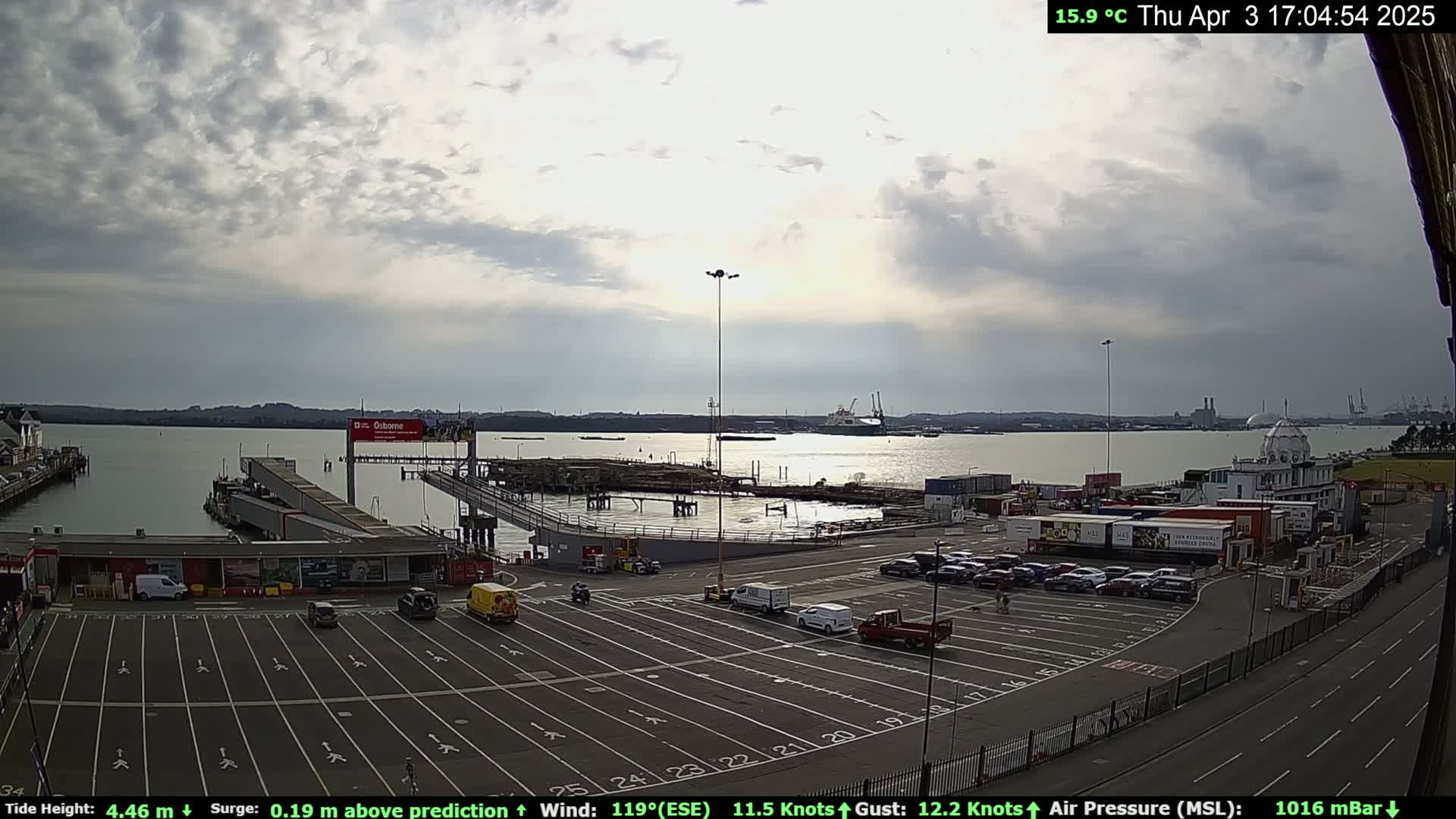 A mostly cloudy day at a ferry terminal shows a large body of water, several parked cars, and a few buildings with containers nearby.