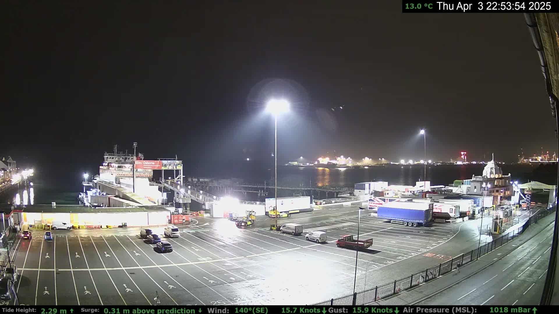 A nighttime view of a ferry terminal with several vehicles parked in the lot, a ferry docked, and other vessels visible across the water under a clear, dark sky.