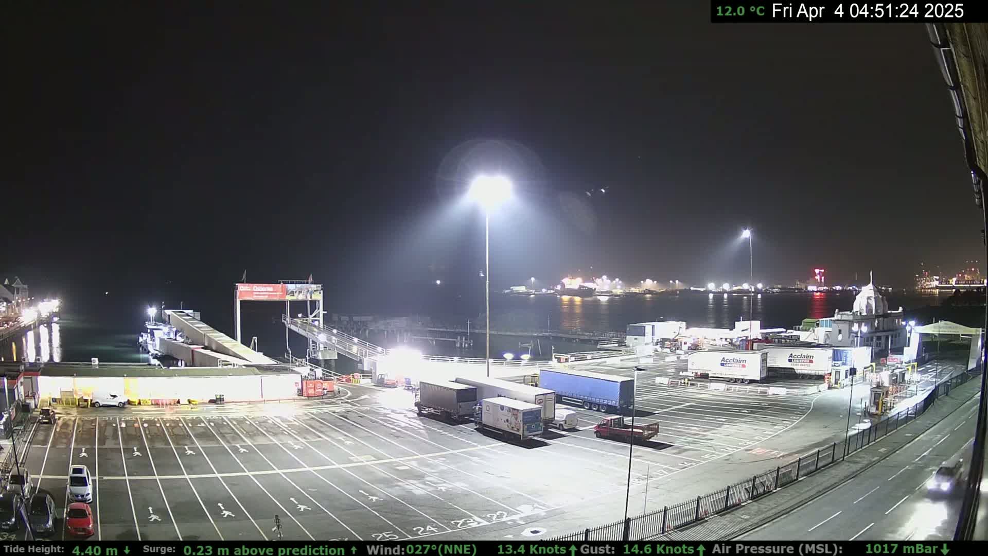 A nighttime view of a ferry terminal with several trucks parked in the lot, a ferry docked at the terminal, and a body of water in the background under a clear sky.