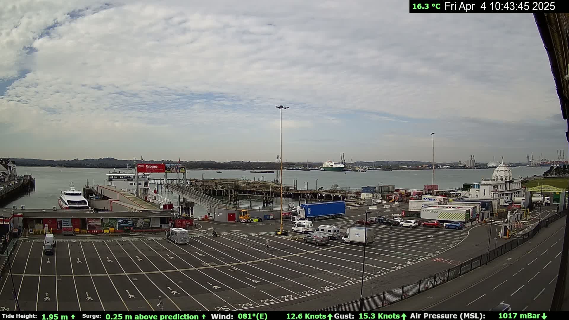 An overcast day at a ferry terminal shows a mostly empty parking lot with several vehicles, a ferry docked at the pier, and a large body of water with cargo ships in the distance.