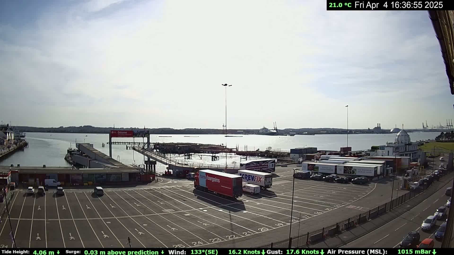 A sunny day at a harbor shows a ferry terminal with several vehicles parked in the lot and several semi-trucks at the docks, alongside calm water.