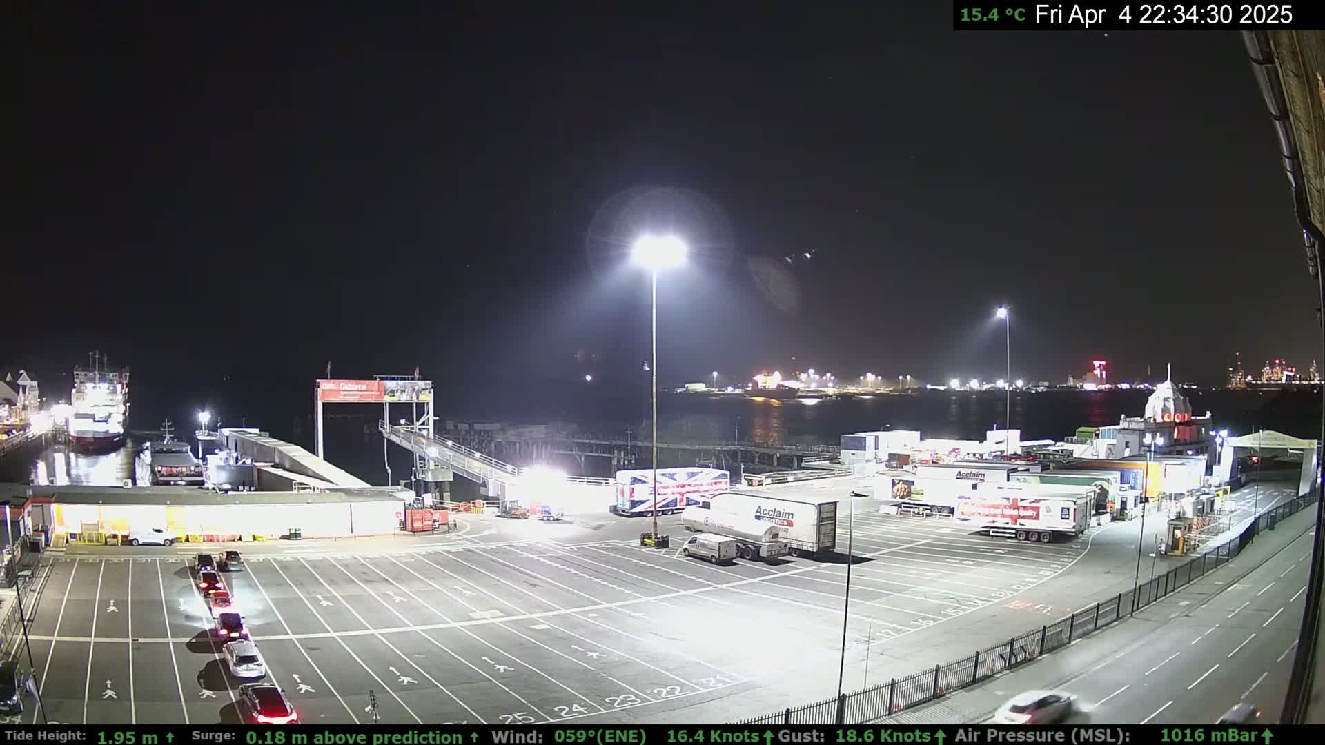 A nighttime view of a ferry terminal with several vehicles parked and a ferry docked, under a clear, dark sky.