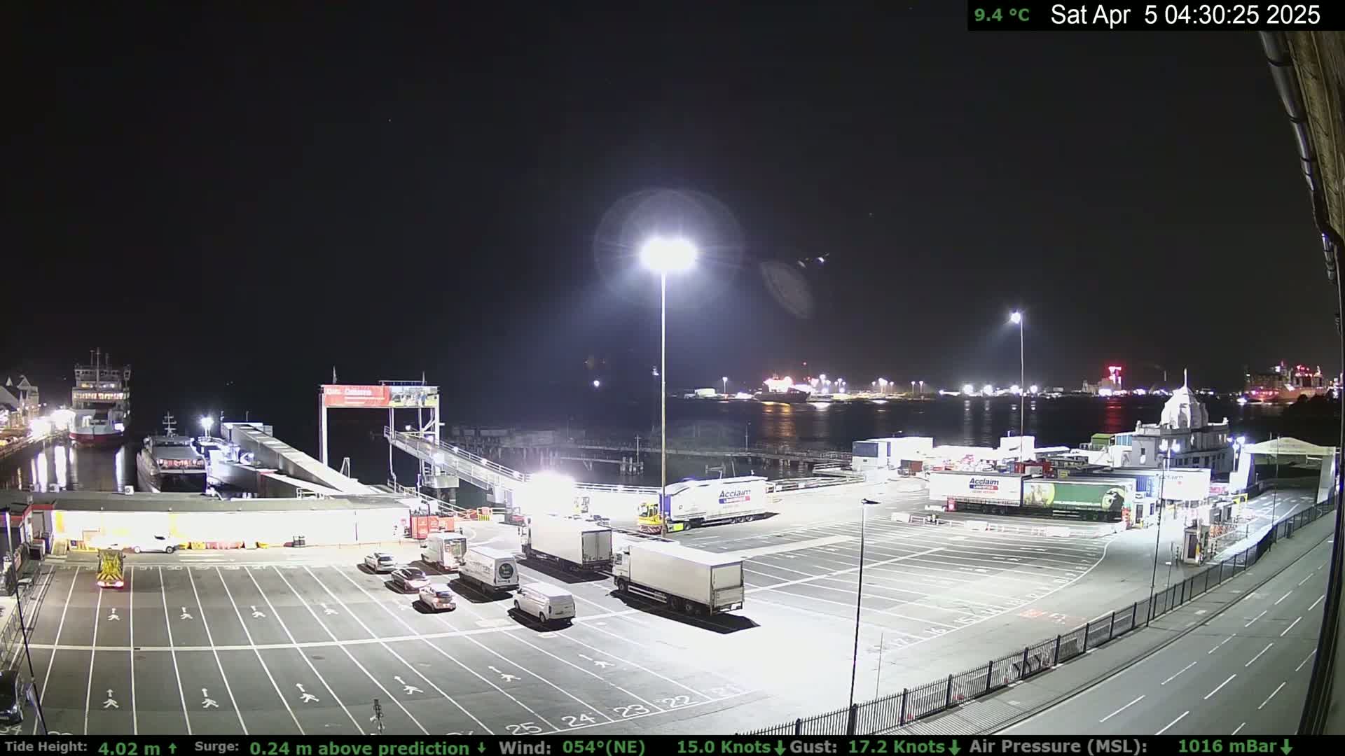 A nighttime view of a ferry terminal with several trucks parked in a lot, illuminated by bright lights, under a clear, cool night sky.