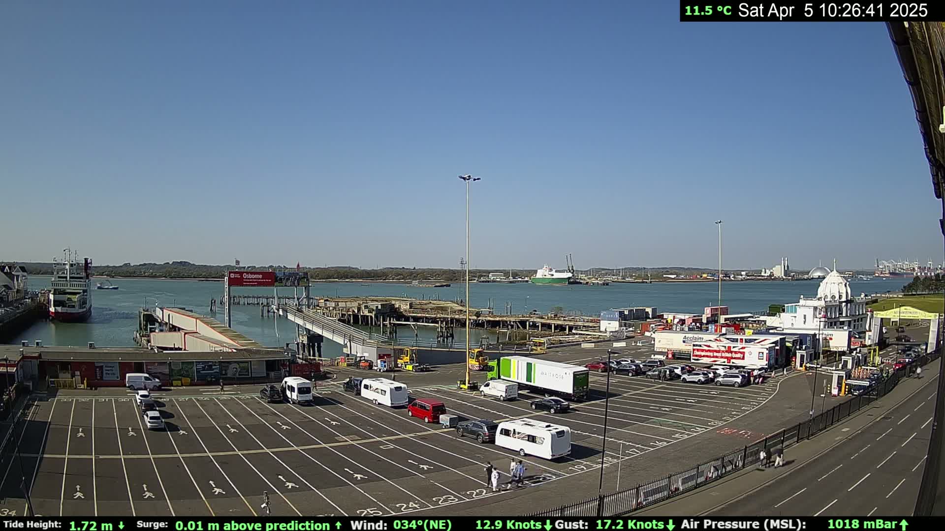 A ferry terminal with several vehicles parked in a lot, a ferry docked, and calm waters under a clear blue sky.