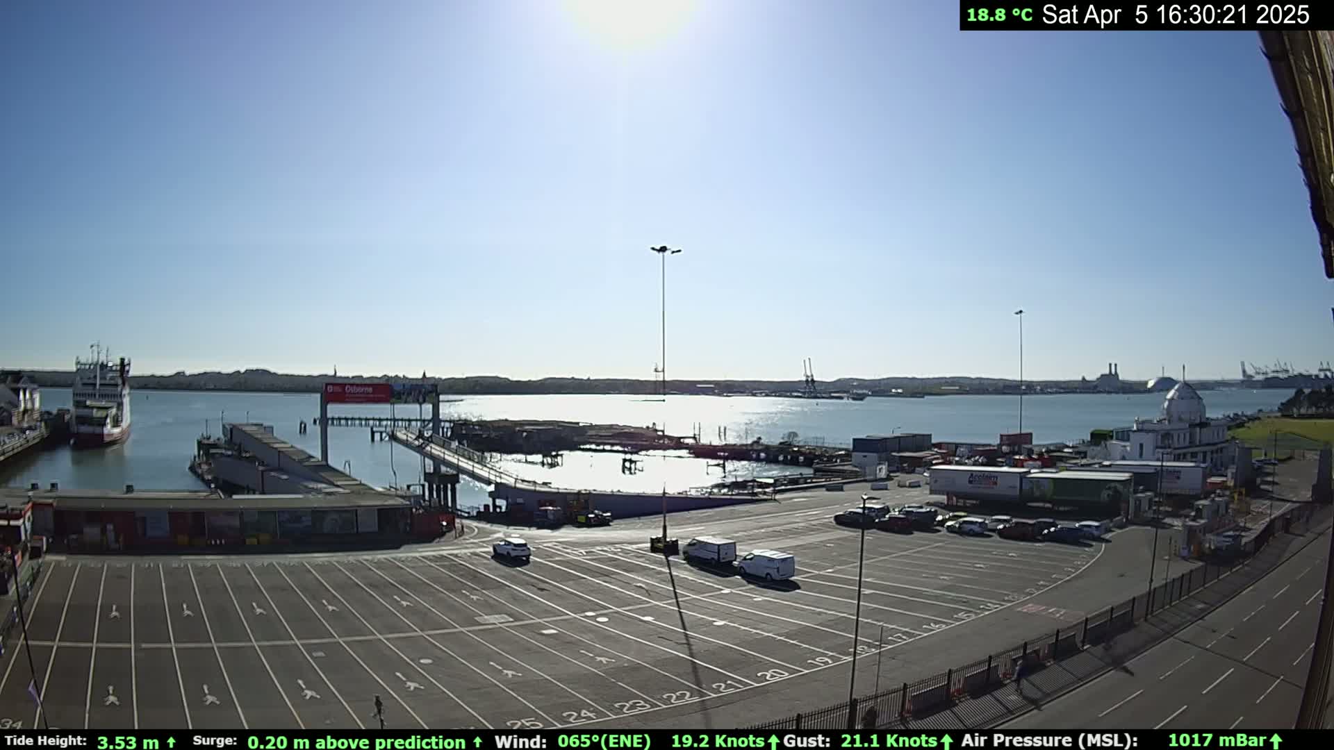 A sunny waterfront scene shows a ferry terminal with a mostly empty parking lot, several docked ships, and calm water under a clear blue sky.