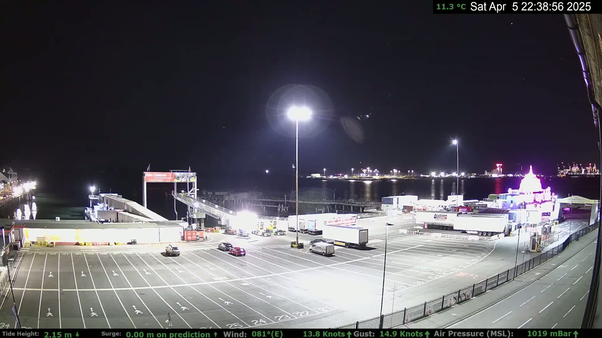 A nighttime view of a ferry terminal with several vehicles parked in the lot, a ferry boat in the water, and city lights in the background under a clear, starry sky.