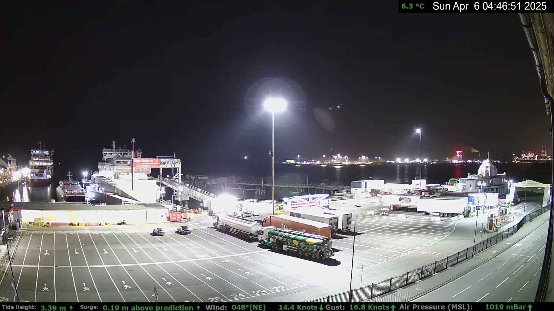 A nighttime view of a ferry terminal with several vehicles parked, two ferries docked, and calm water.