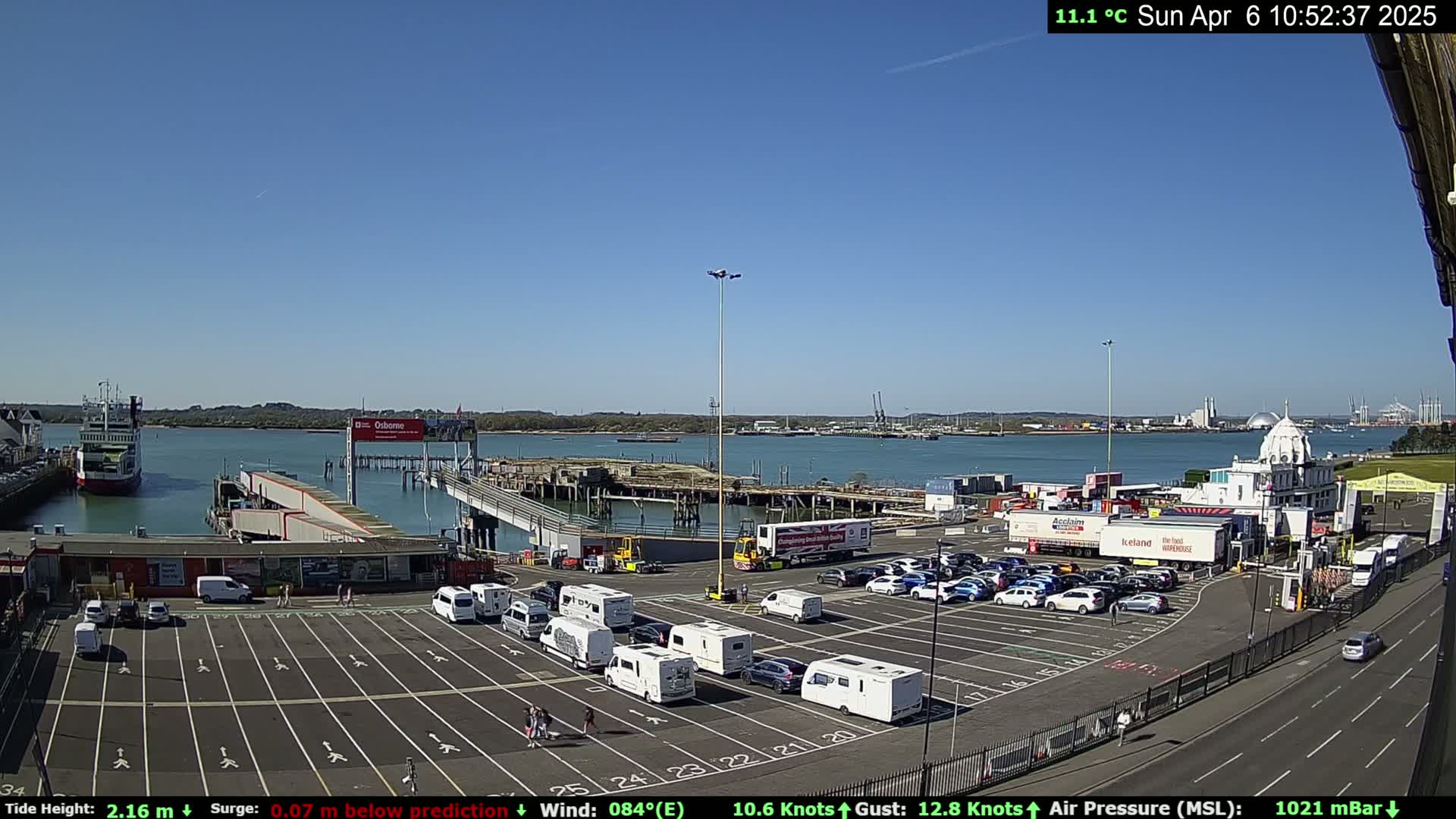 A ferry terminal with a large parking lot full of camper vans and cars, under a clear blue sky, is shown on a sunny day.