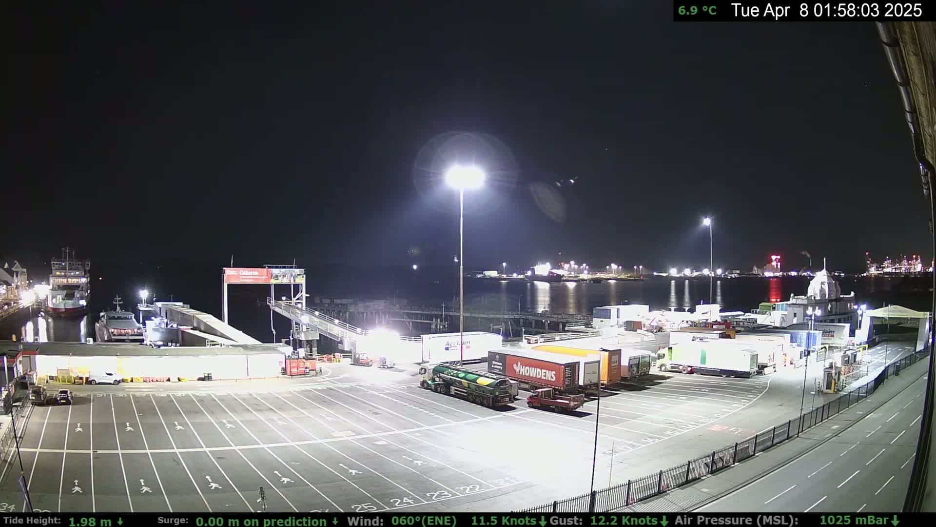 A nighttime view of a ferry terminal with several trucks and ferries docked, under a clear sky.