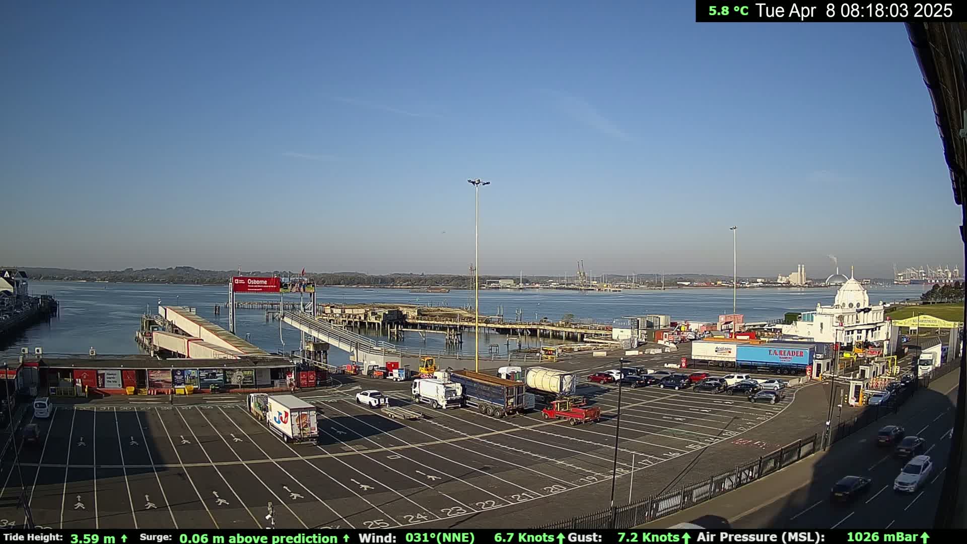 A ferry terminal with a large parking lot and several trucks, situated on a calm body of water under a clear blue sky.