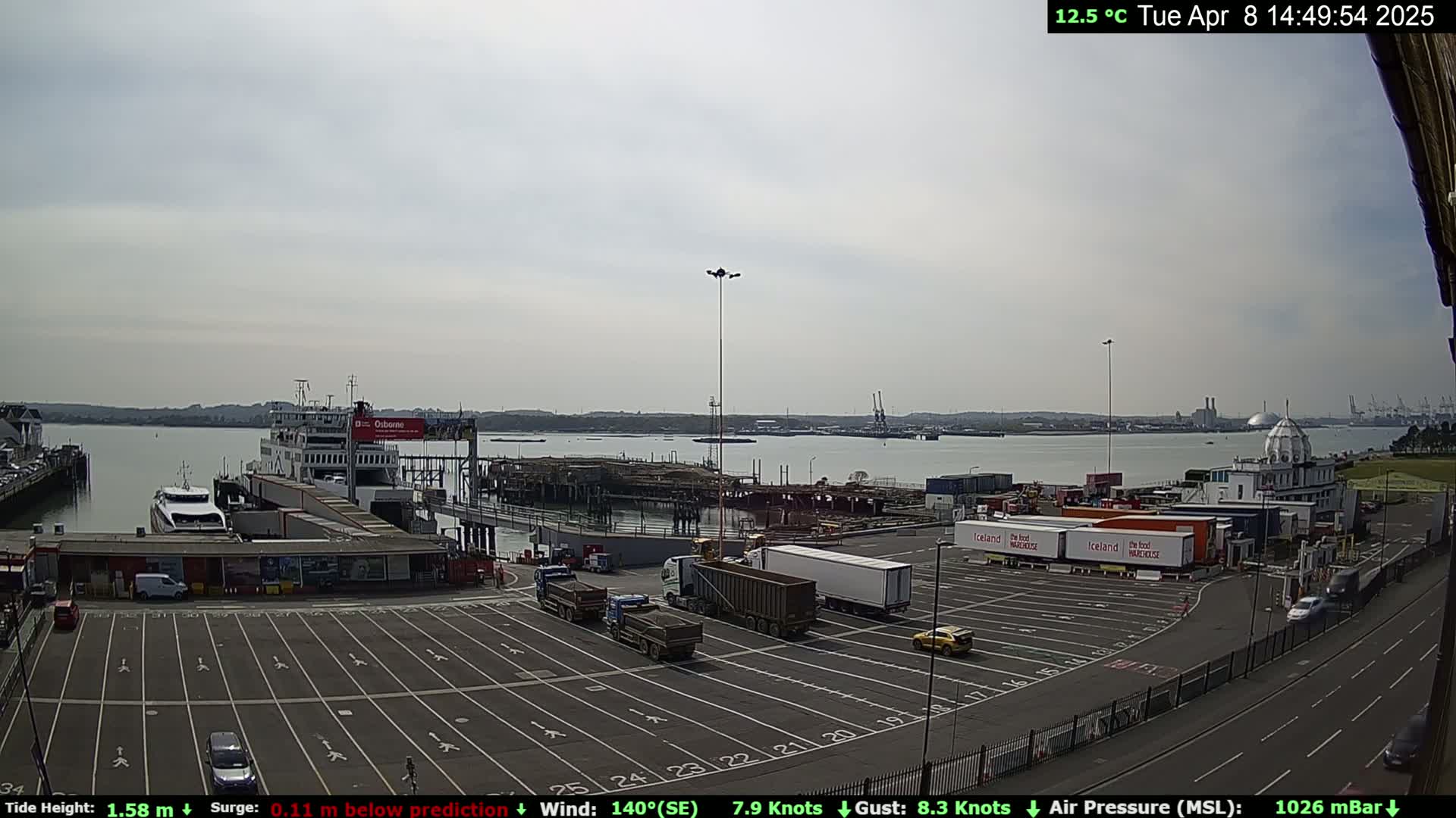 A ferry is docked at a port on a partly cloudy day, with several trucks and cars in the parking lot, and a body of water visible in the background.