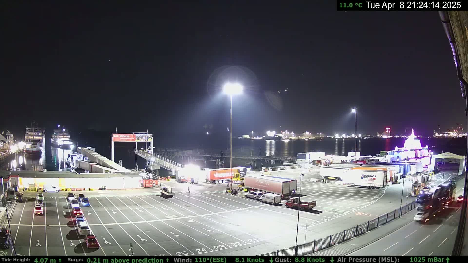A nighttime view of a ferry terminal with several parked cars and trucks, illuminated by bright lights, under a clear sky.