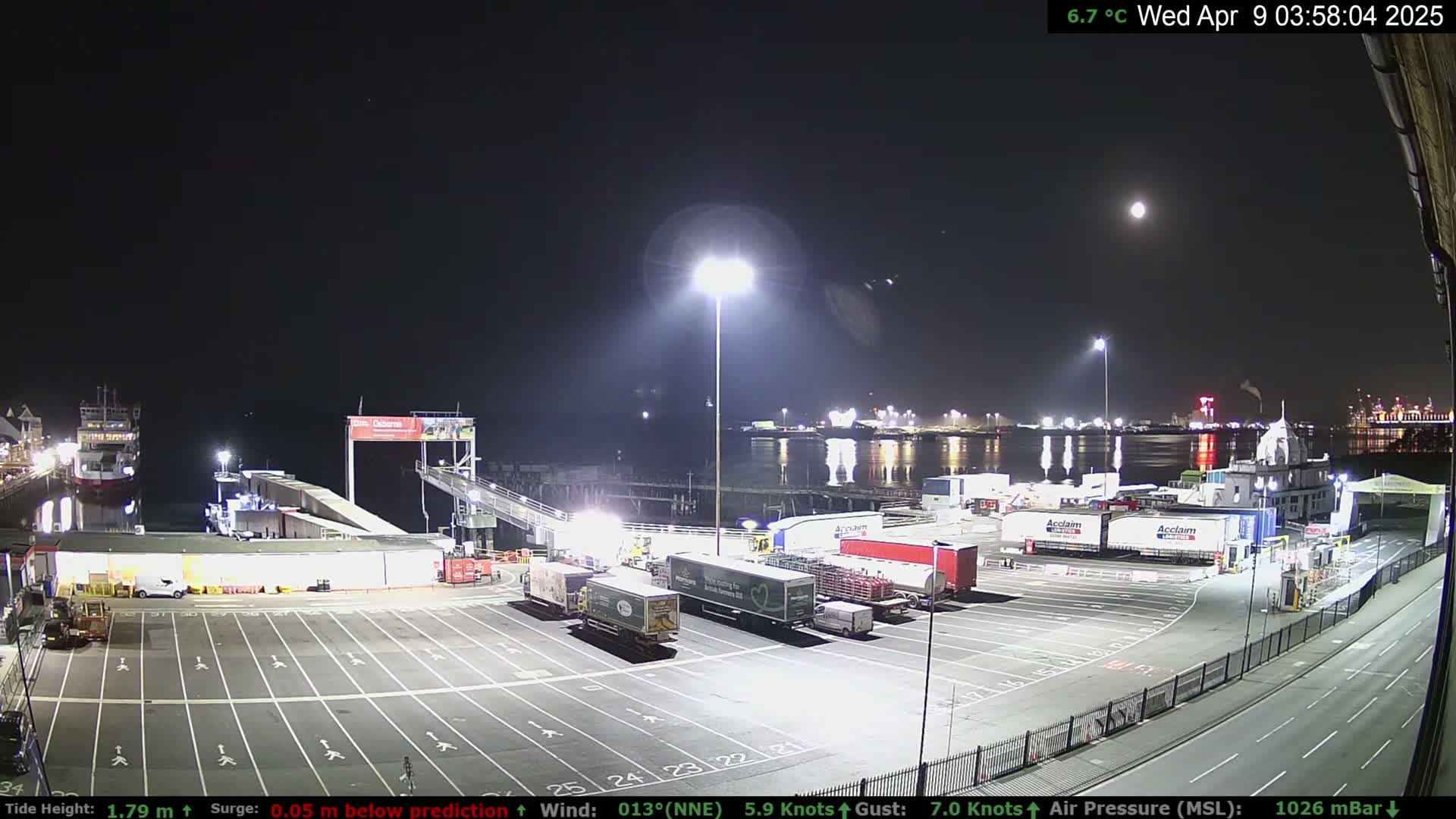 A nighttime view of a ferry terminal with several trucks parked in a lot, a ferry docked, and a calm body of water reflecting lights under a bright moon.