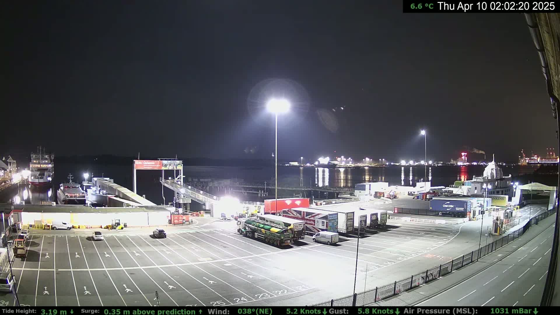 A nighttime view of a ferry terminal with several trucks and cars parked in a lot, along with several ships docked at the water's edge under a clear sky.