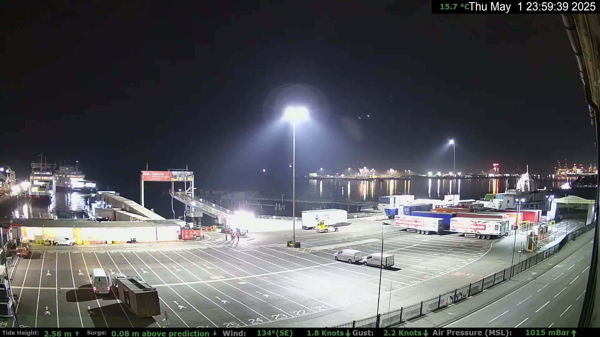 A nighttime view of a ferry terminal with several trucks and cars parked in a lot,  ferries moored at the dock, and calm water reflecting lights across the harbor.