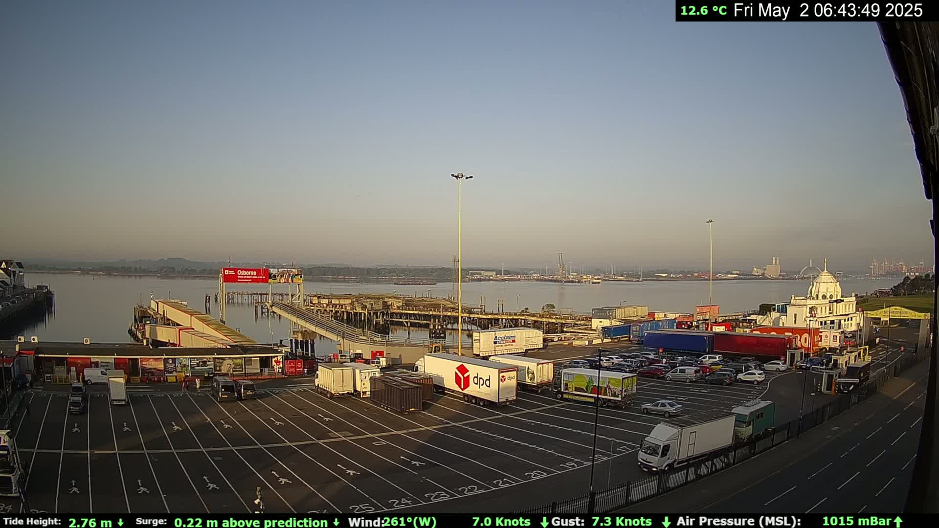A ferry terminal with numerous parked trucks and cars is situated on a calm body of water under a clear, light-blue sky.