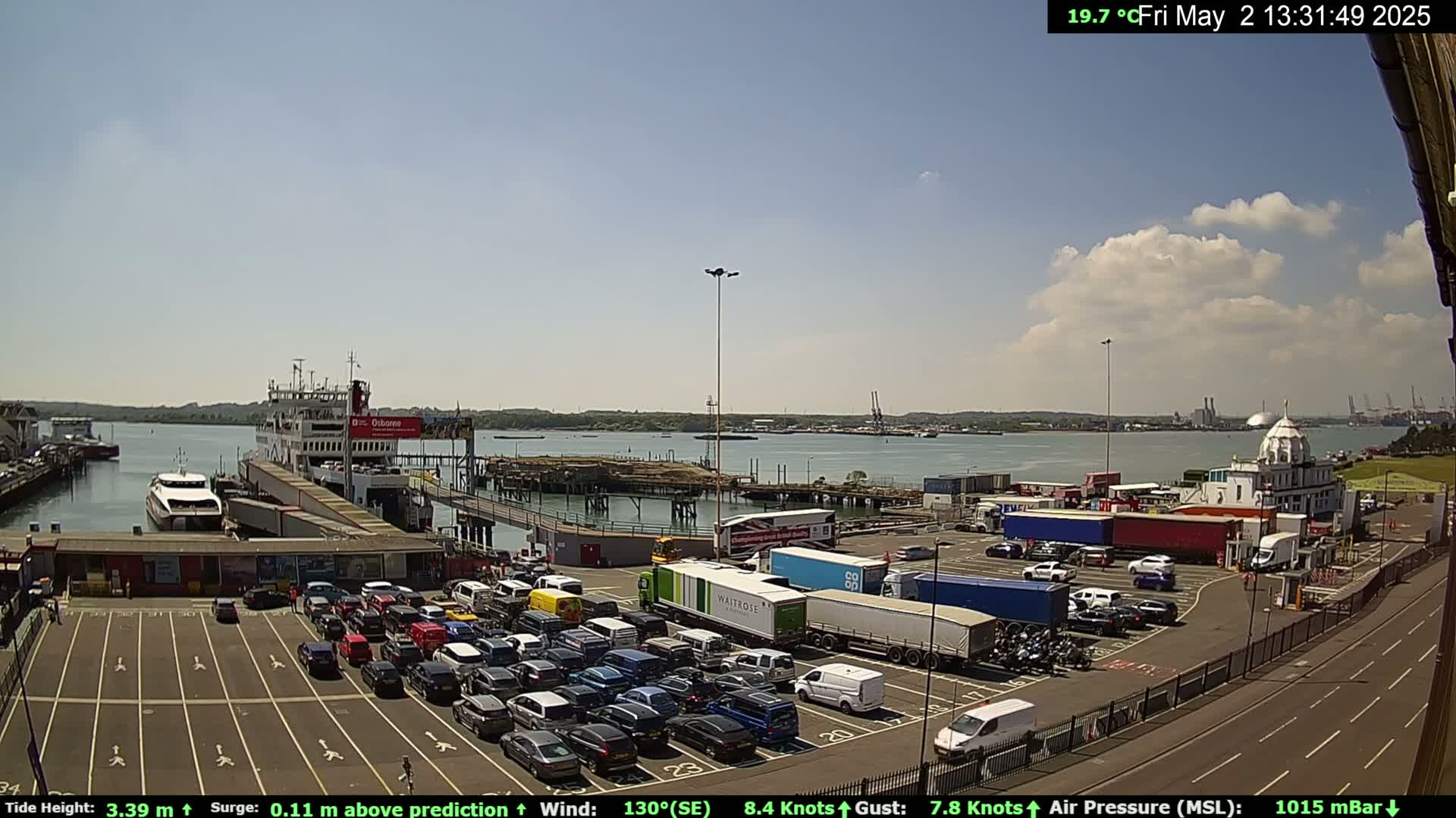 A ferry is docked at a busy port under a partly cloudy sky, with numerous cars and trucks in a large parking lot.