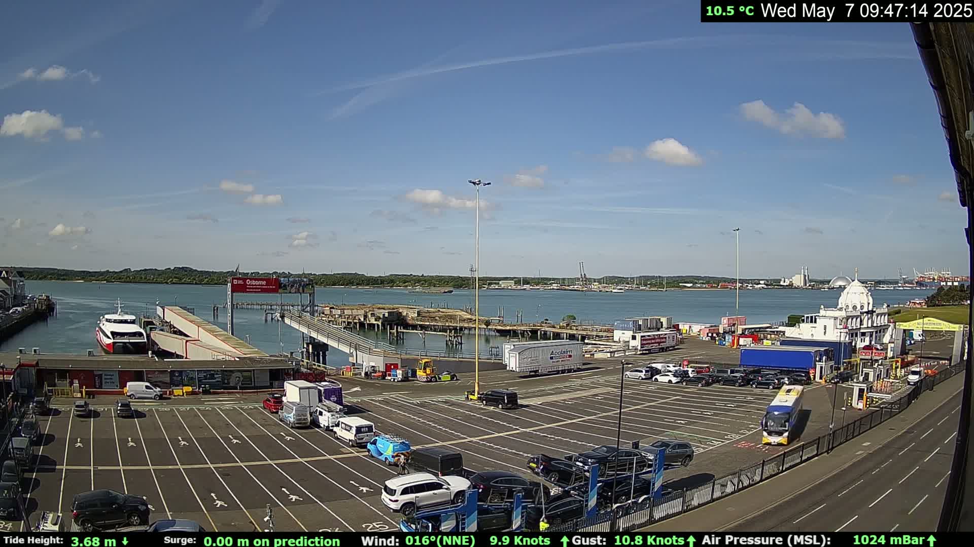 A ferry is docked at a terminal in a sunny harbor, with cars parked in a large lot and buildings surrounding the area.