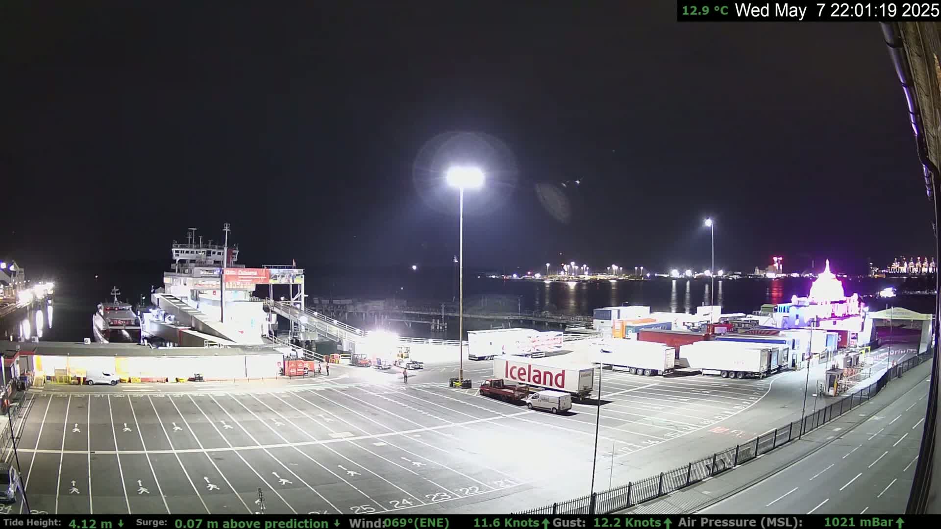 A ferry is docked at a terminal at night, with numerous semi-trailer trucks parked nearby under bright lights.