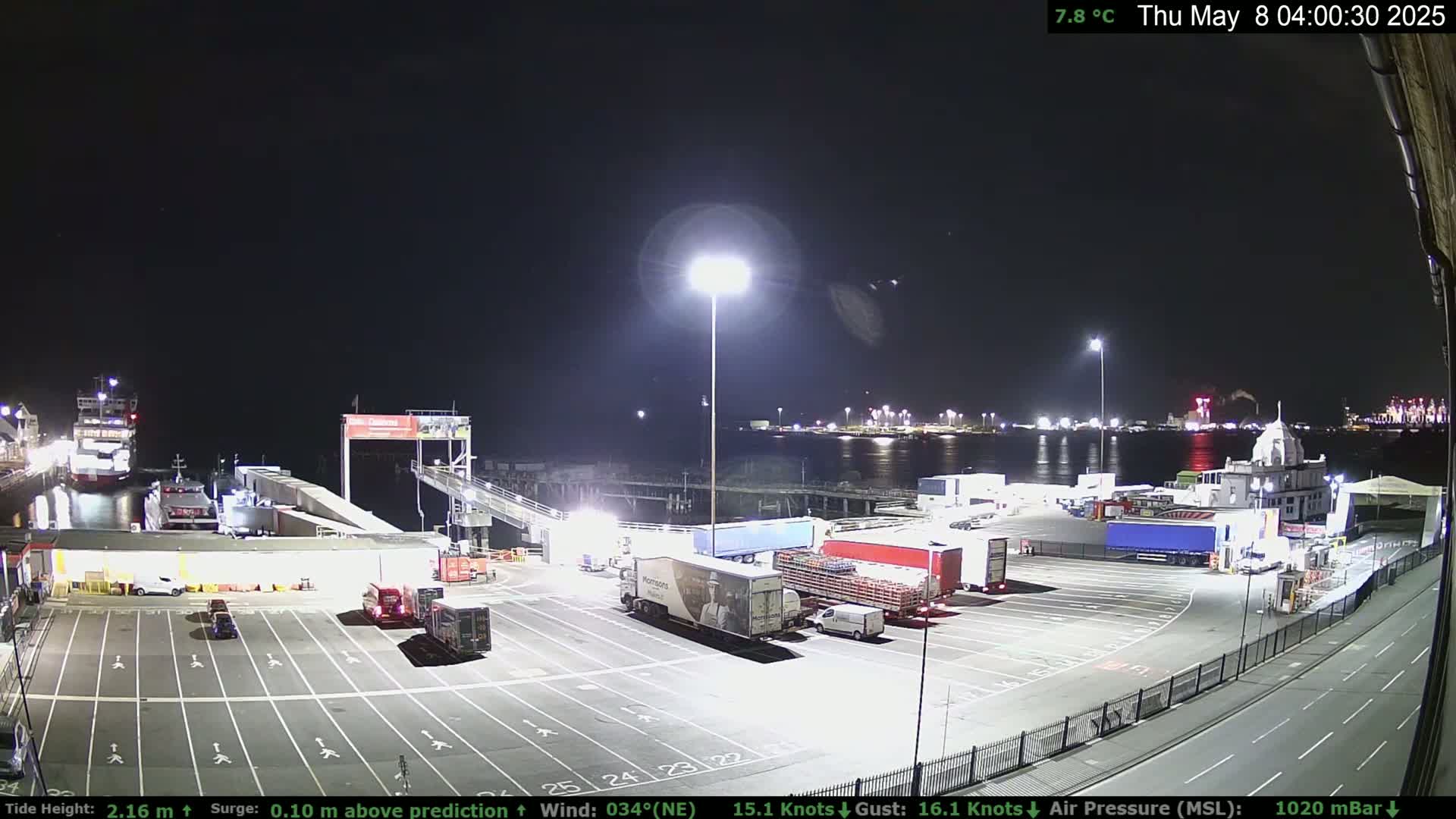 A nighttime view of a ferry terminal with several trucks parked in the lot and ferries docked, under a clear, cool sky.