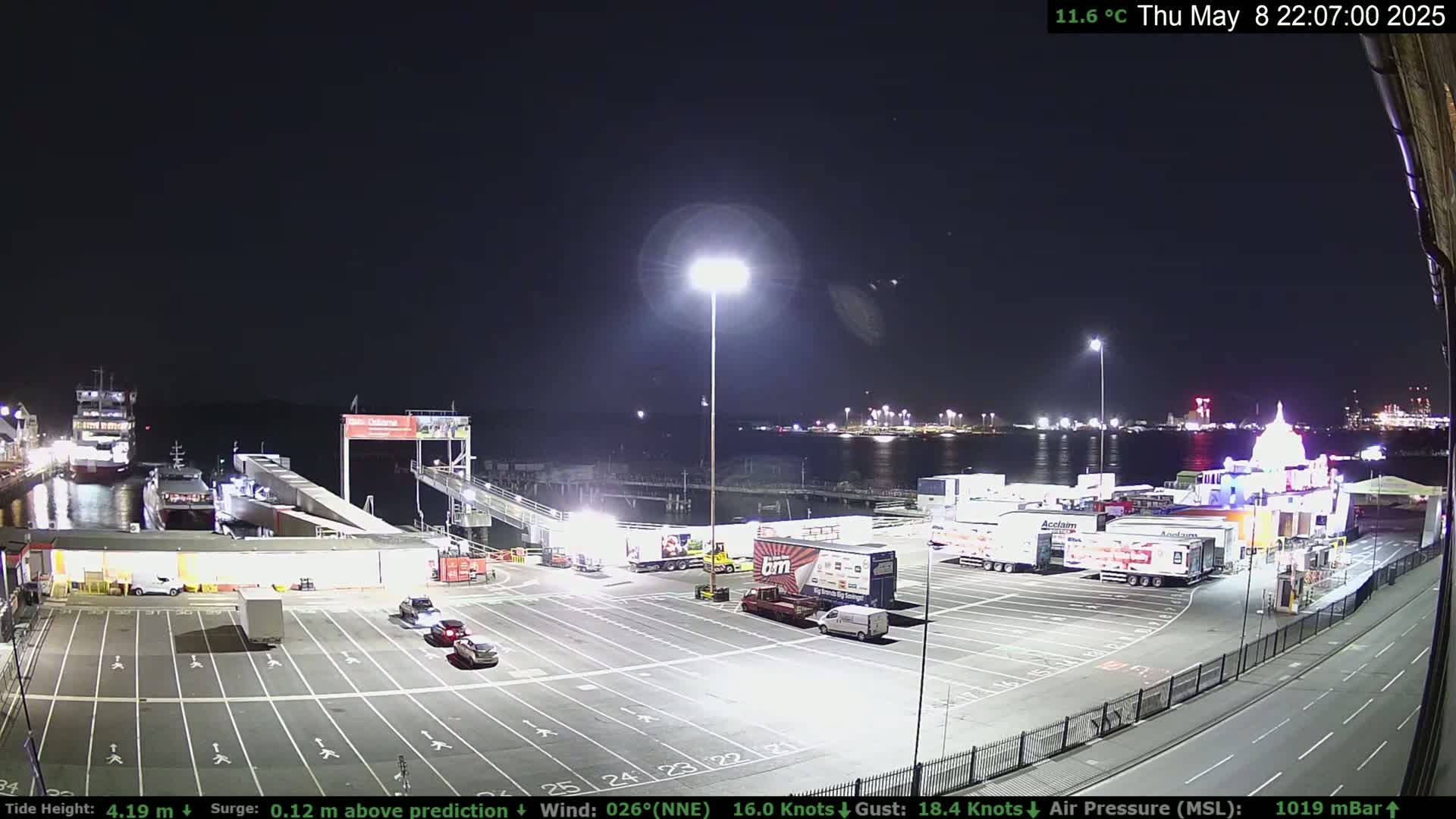 A nighttime view of a ferry terminal with several vehicles parked, a ferry docked, and a brightly lit building in the background under a clear, dark sky.