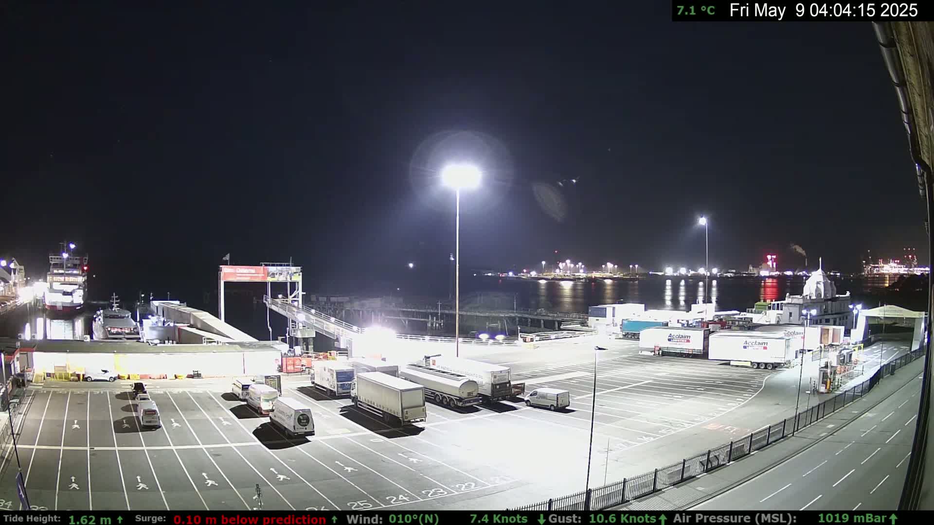 A nighttime view of a ferry terminal with several large trucks parked in a lot, a ferry docked, and lights illuminating the scene.