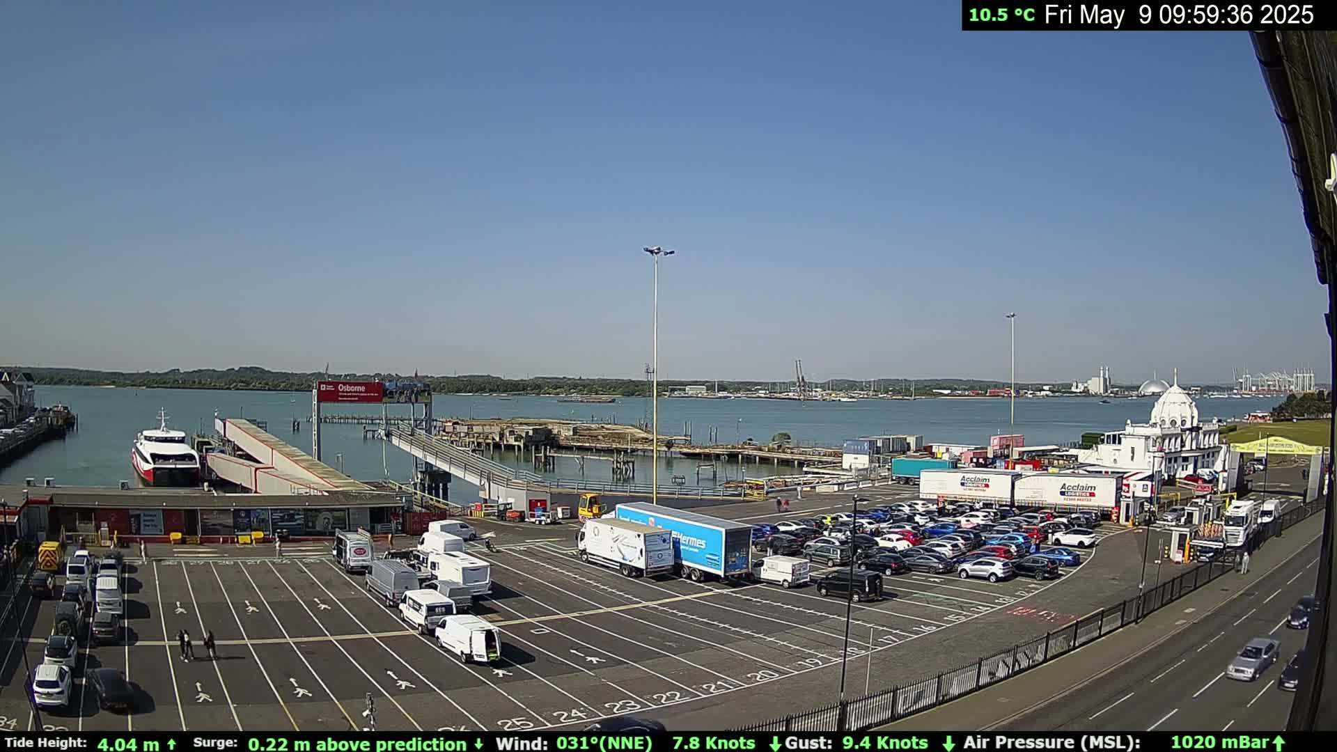 A ferry is docked at a terminal with numerous cars parked in a lot, under a clear blue sky.