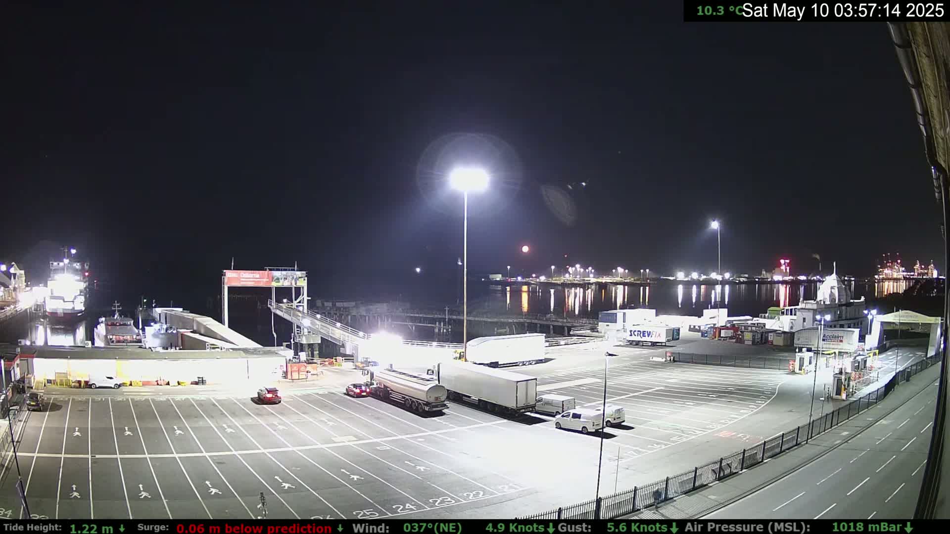 A nighttime view of a ferry terminal with several vehicles parked in a lot, a ferry docked, and a red moon visible in the distance under a clear sky.