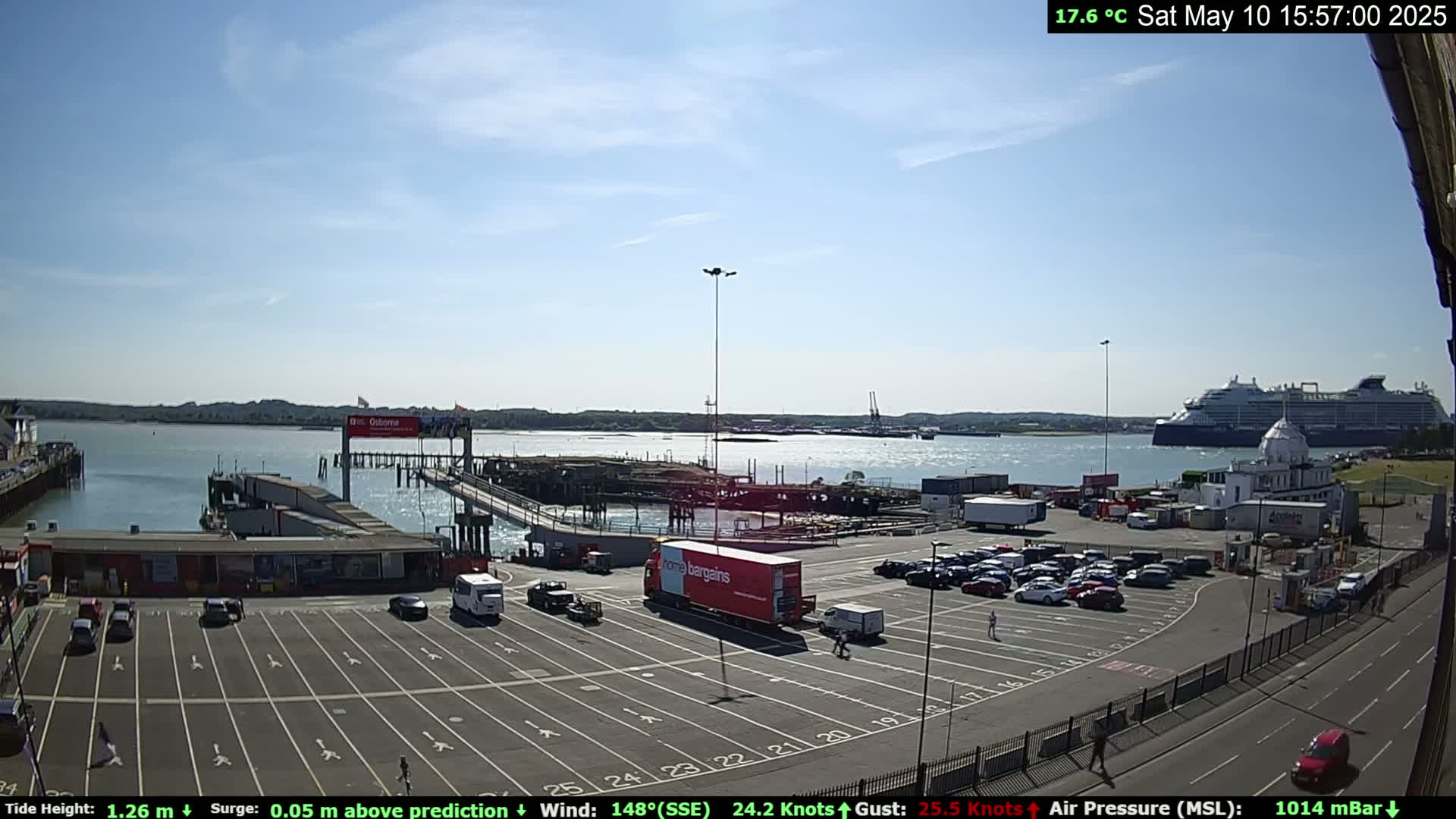 A sunny day at a port shows a large cruise ship in the water, a parking lot full of cars, and several buildings near the docks.