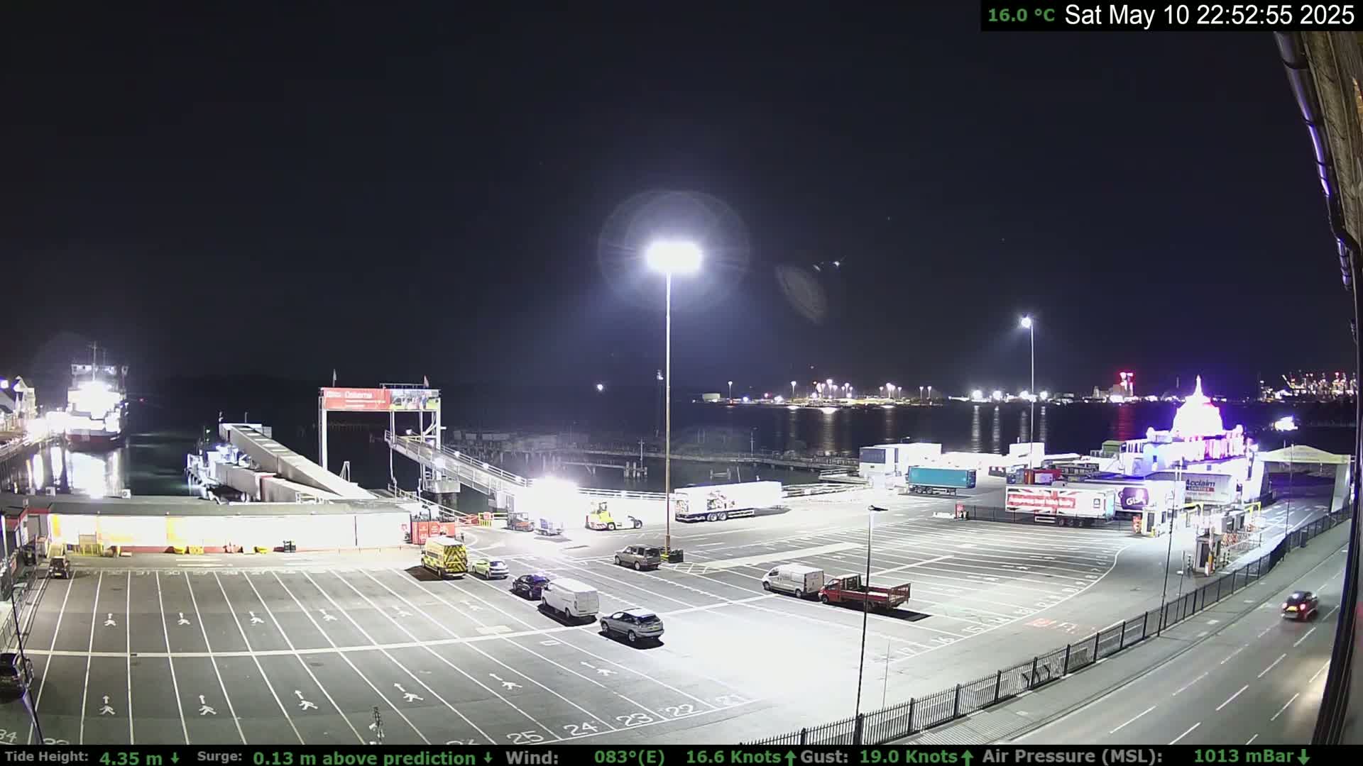 A nighttime view of a ferry terminal with several vehicles in the parking lot and a ferry docked, under a clear sky.