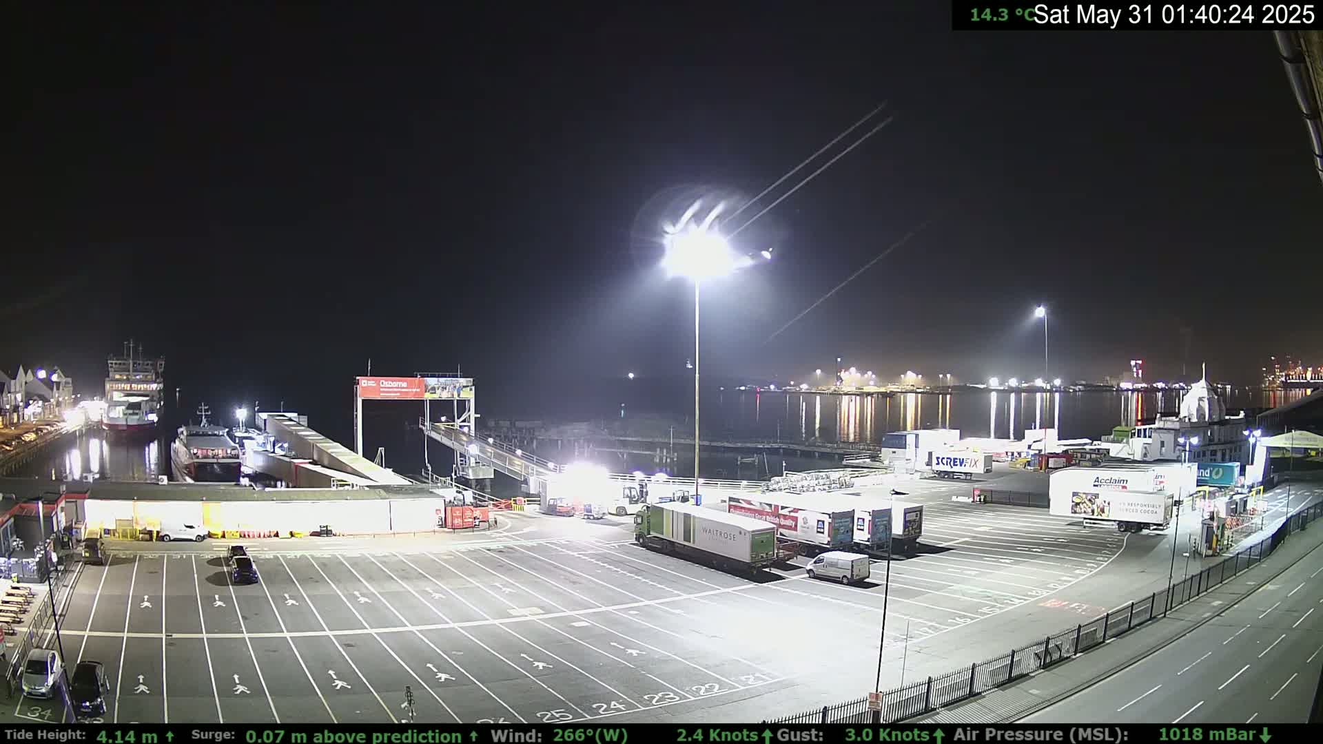 A nighttime view of a ferry terminal with several docked ships, parked trucks, and cars under clear skies.