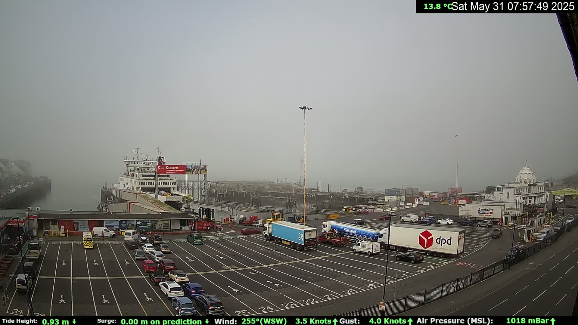 A ferry is docked at a port on a foggy day, with numerous cars and trucks in the parking lot.