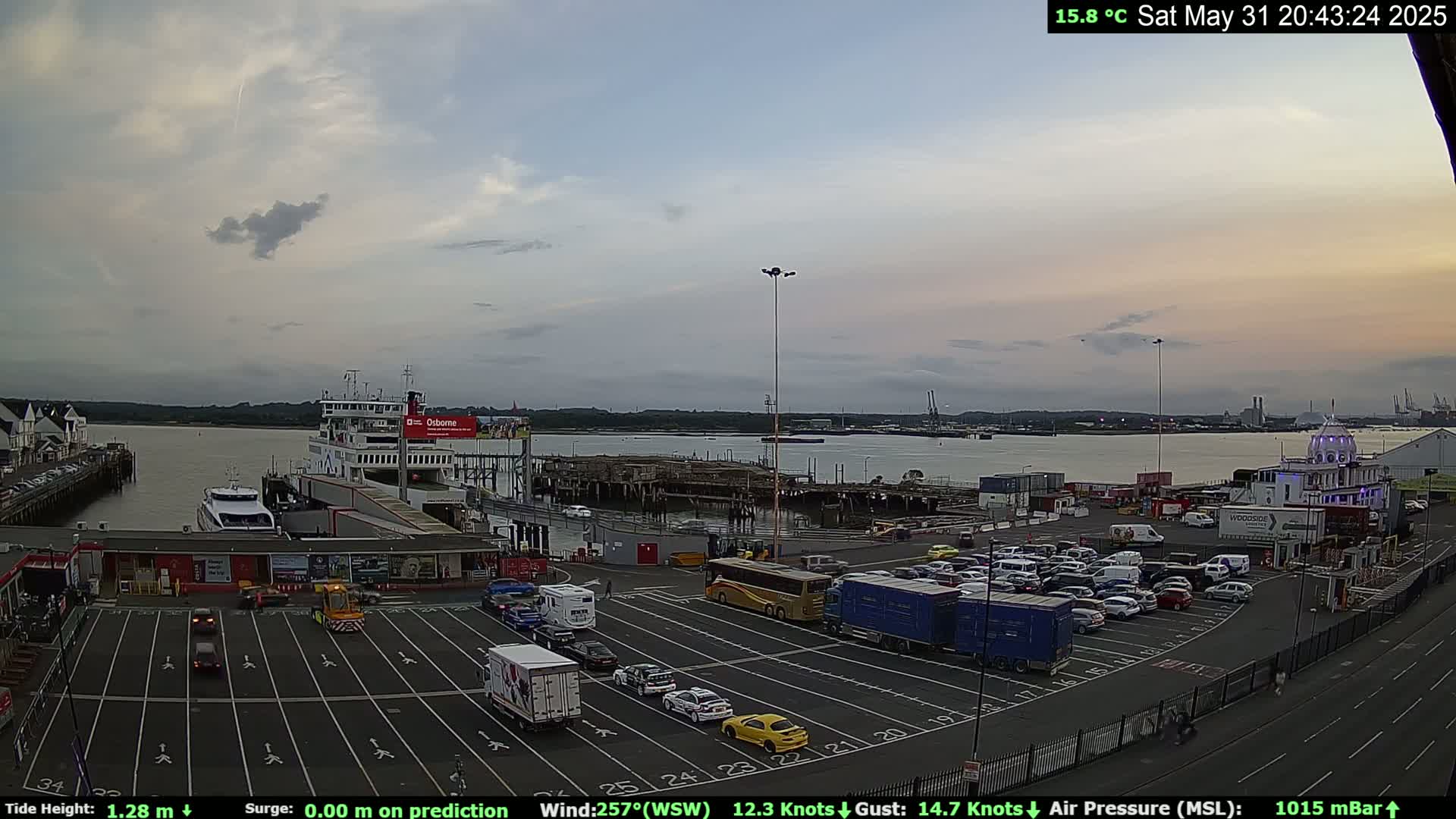 A ferry terminal at dusk, with a mostly cloudy sky, shows numerous cars parked in the lot and a ferry docked at the pier.