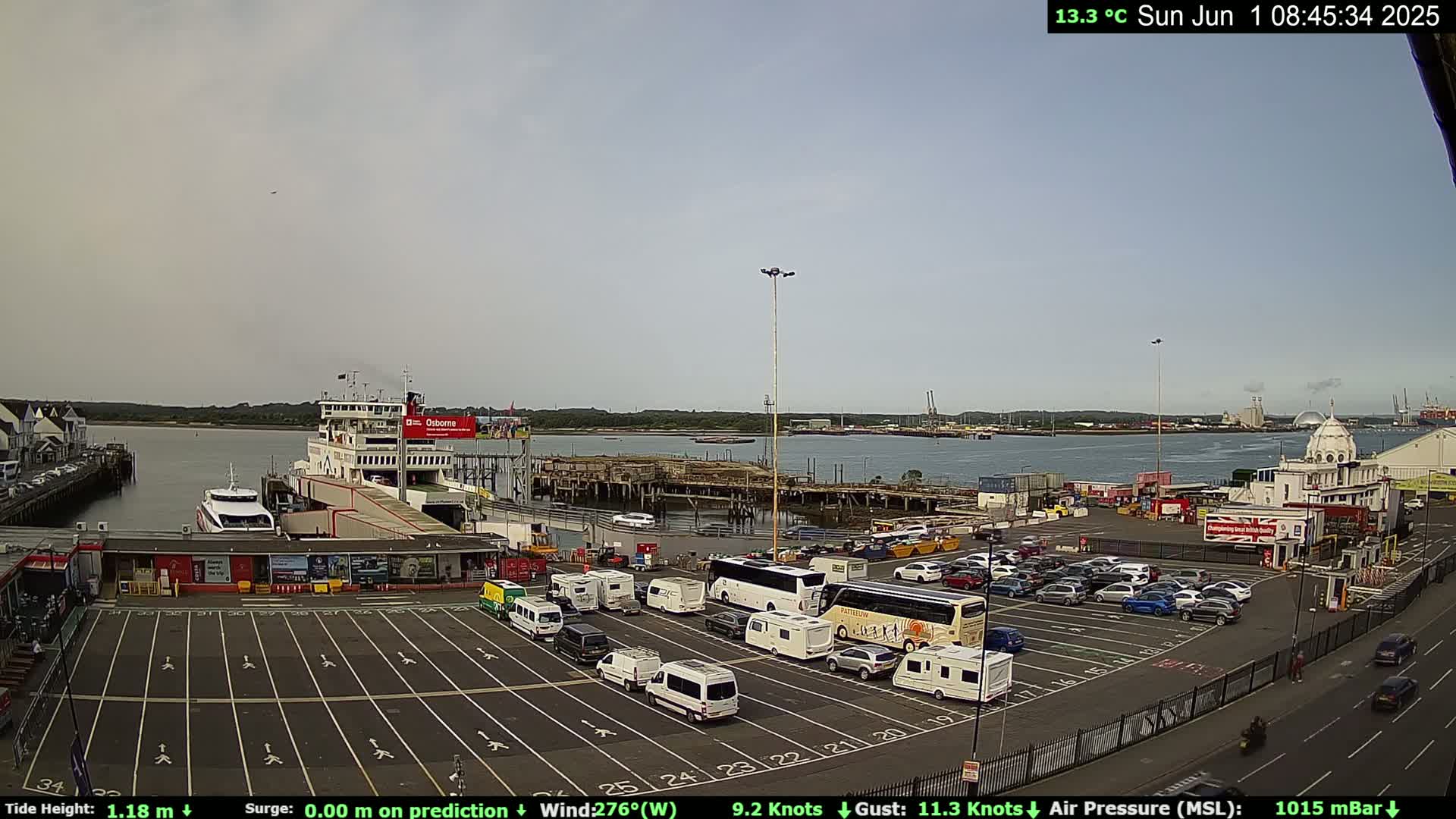 A ferry terminal with a ferry docked, numerous vehicles parked in a lot, and calm water in the background under an overcast sky.