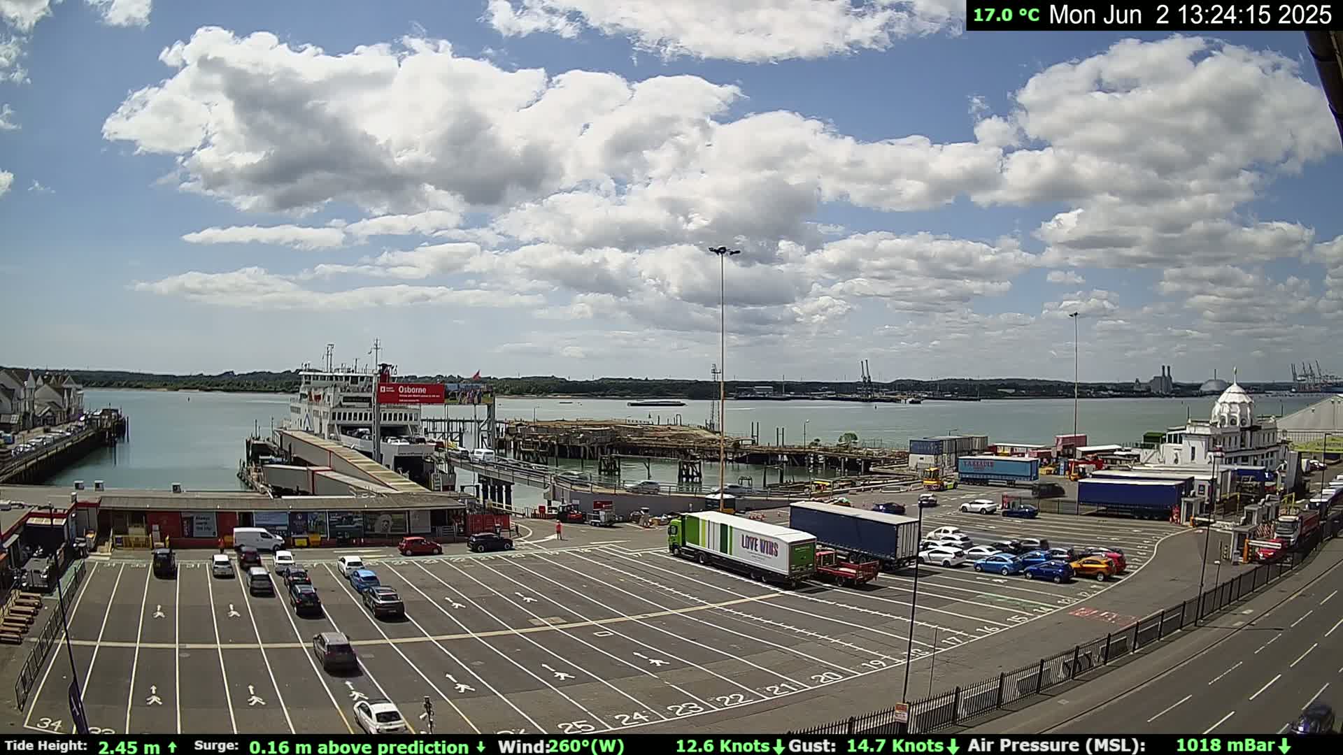 A ferry is docked at a port on a partly cloudy day with a temperature of 17 degrees Celsius; cars are parked in the surrounding lot, and several large trucks are also visible.