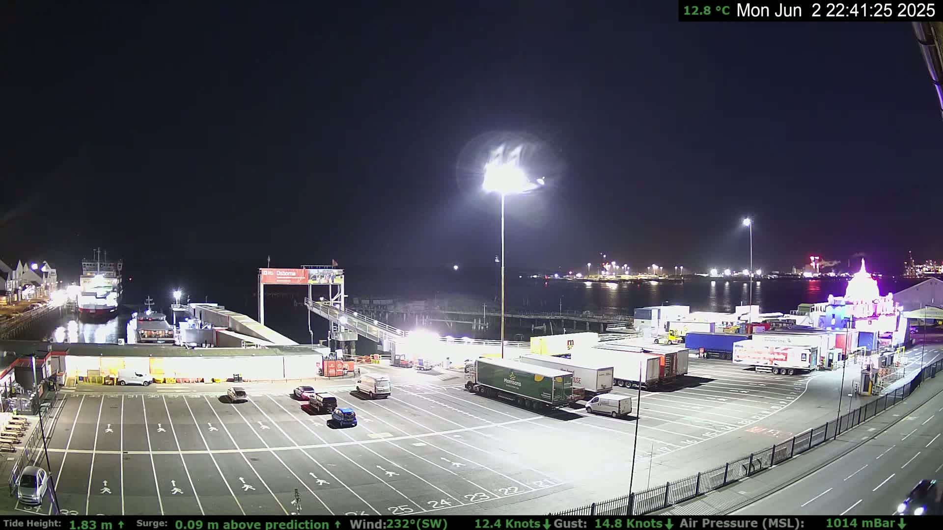 A nighttime view of a ferry terminal with several trucks and cars parked in the lot, a ferry docked, and calm water under a clear, dark sky.