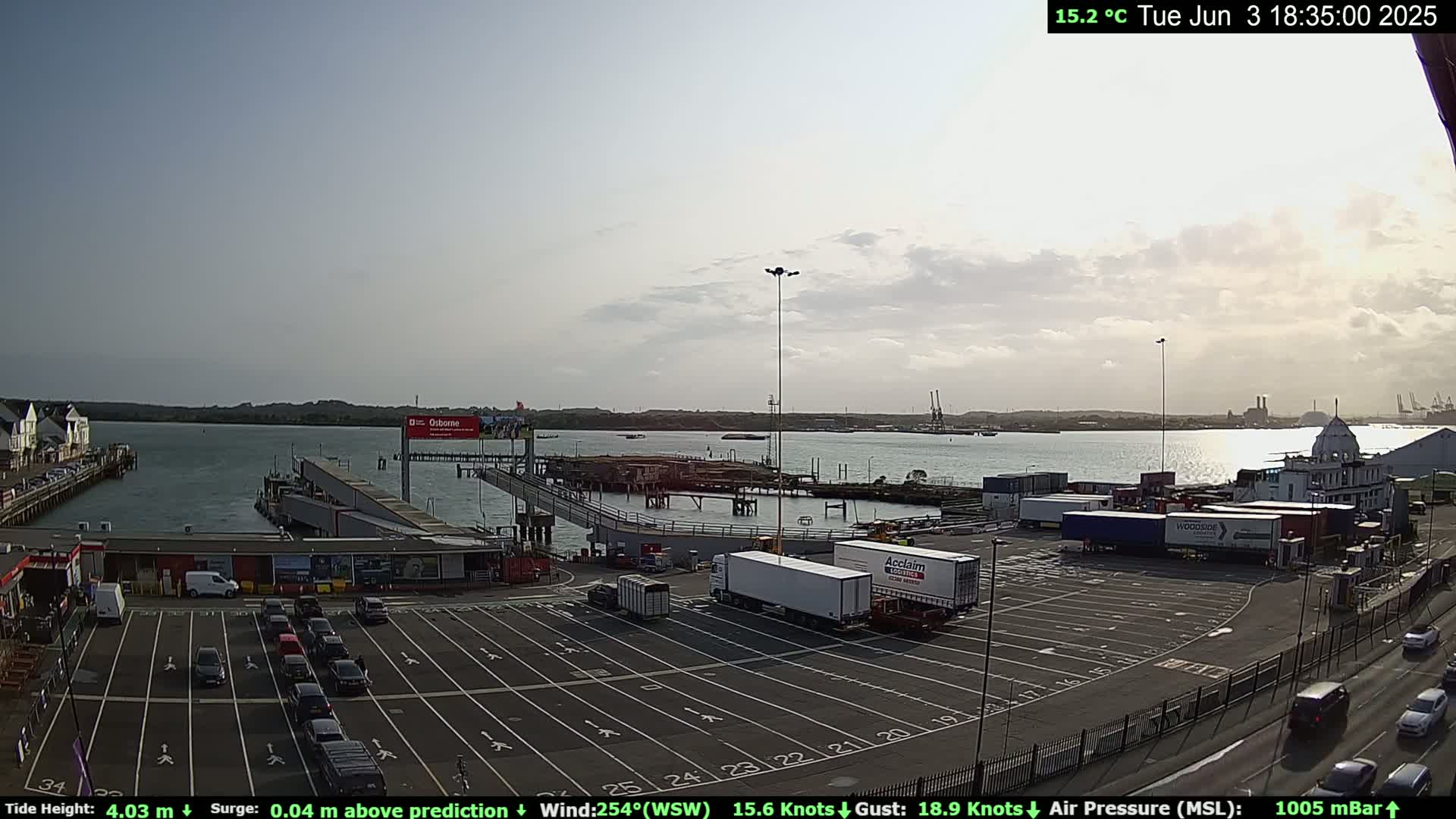 A ferry terminal with a mostly empty parking lot and several large trucks, situated on a body of water under a partly cloudy sky.