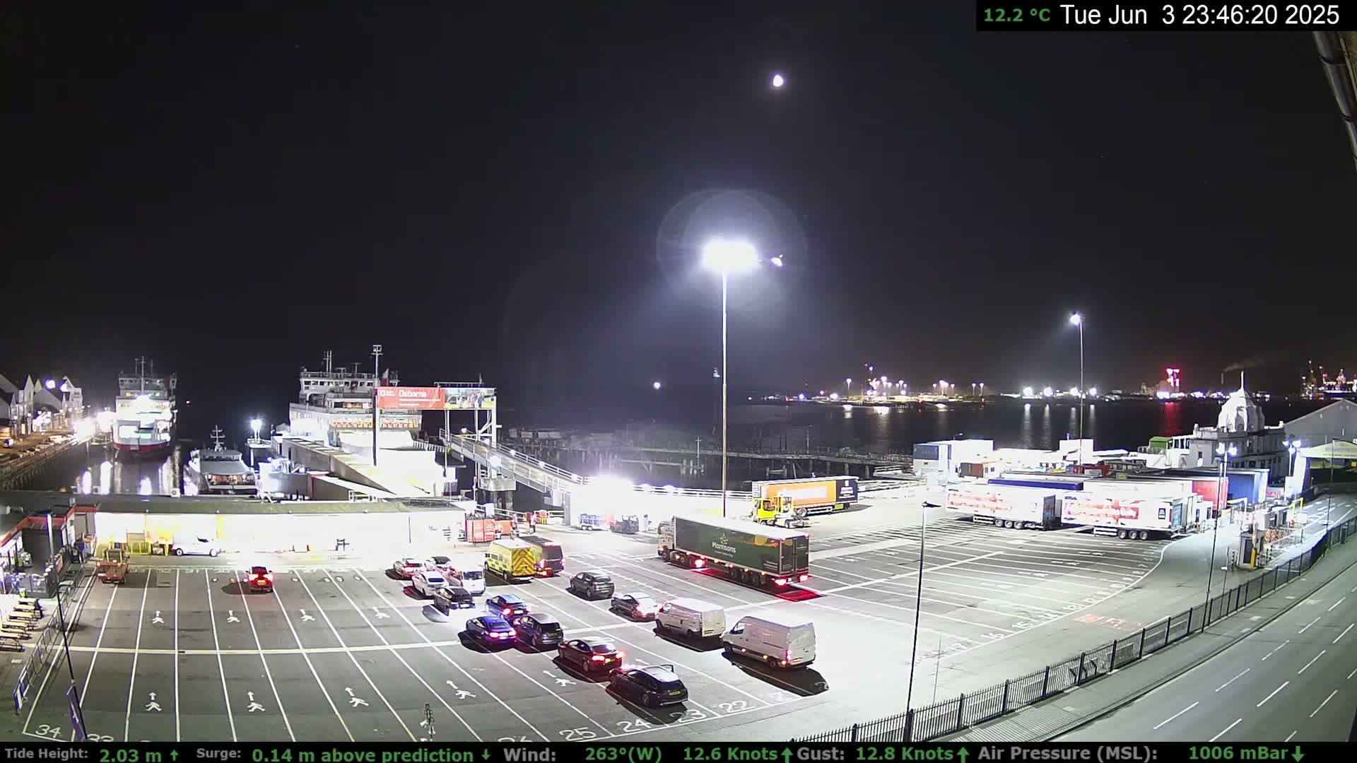 A nighttime view of a ferry terminal with several cars and trucks parked, two ferries docked, and a mostly clear sky with a moon visible.