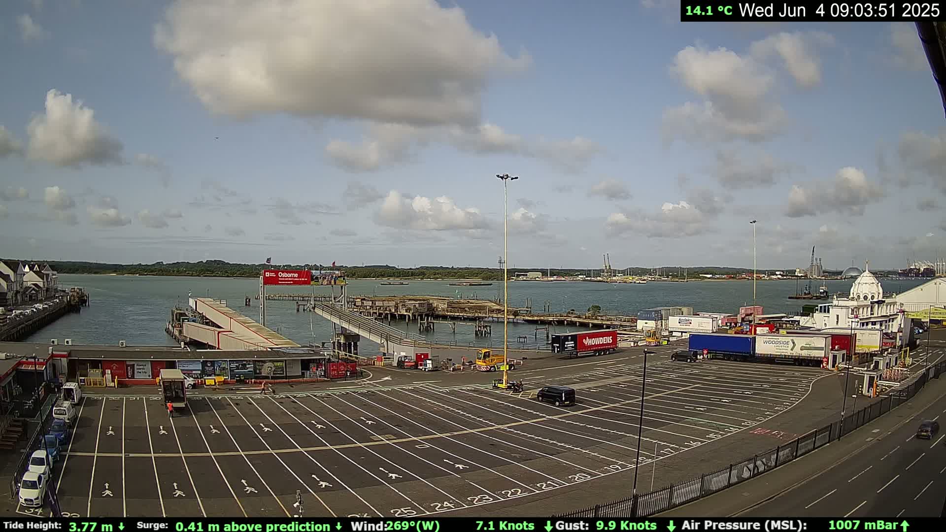 A mostly sunny day at a port with a large empty parking lot, a ferry terminal, and many shipping containers.