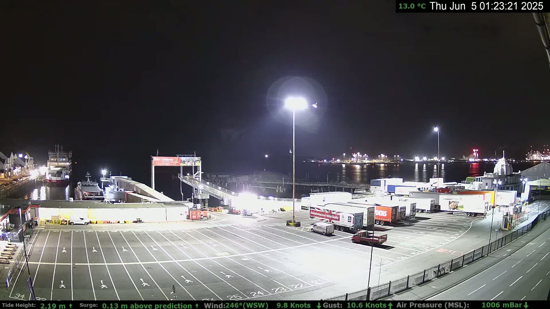 A nighttime view of a ferry terminal with several docked ships, trucks in a parking lot, and calm water.