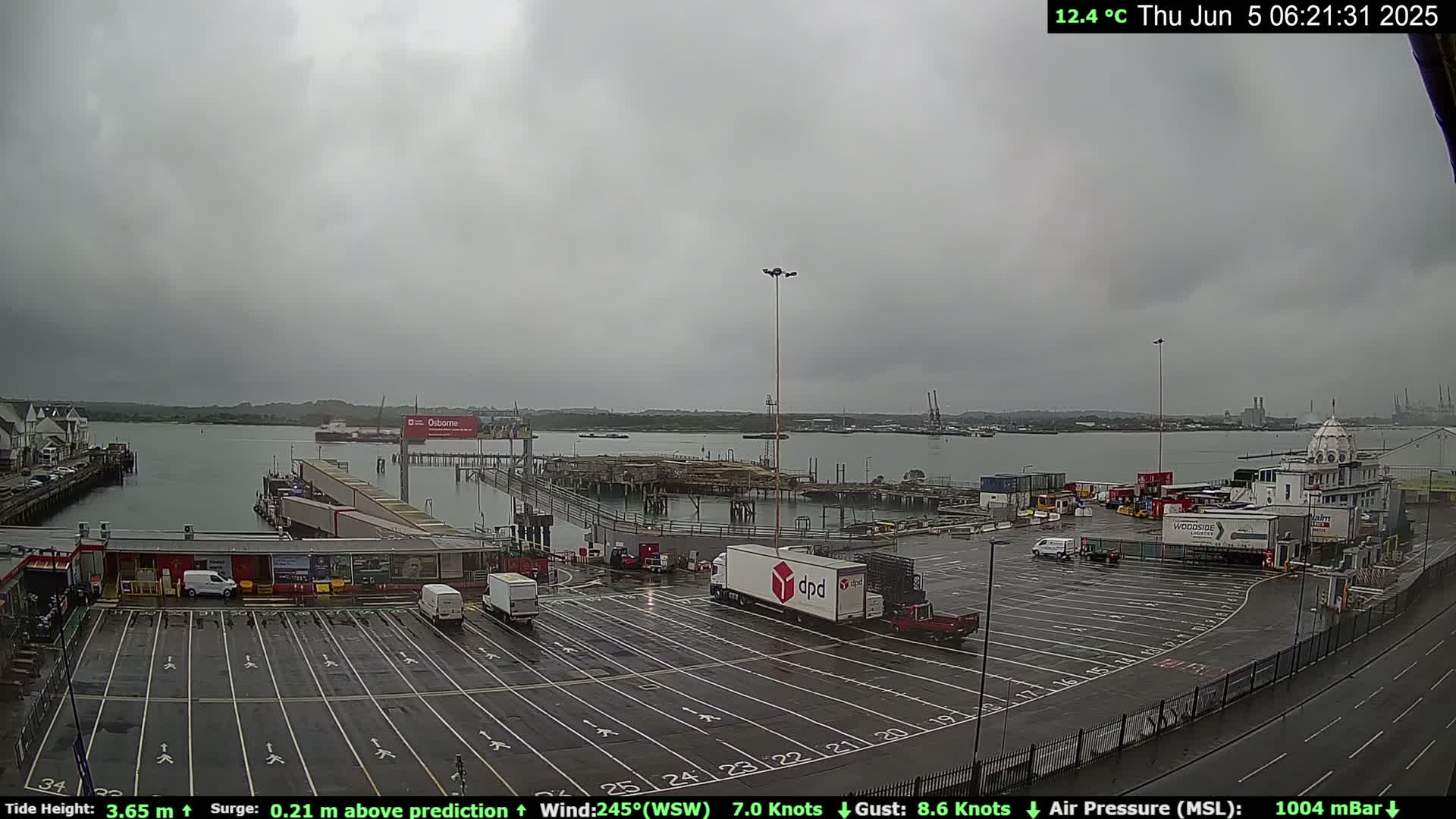 A mostly cloudy, overcast day at a port shows a large empty parking lot with several trucks and vans, a body of water with ships, and buildings in the background.