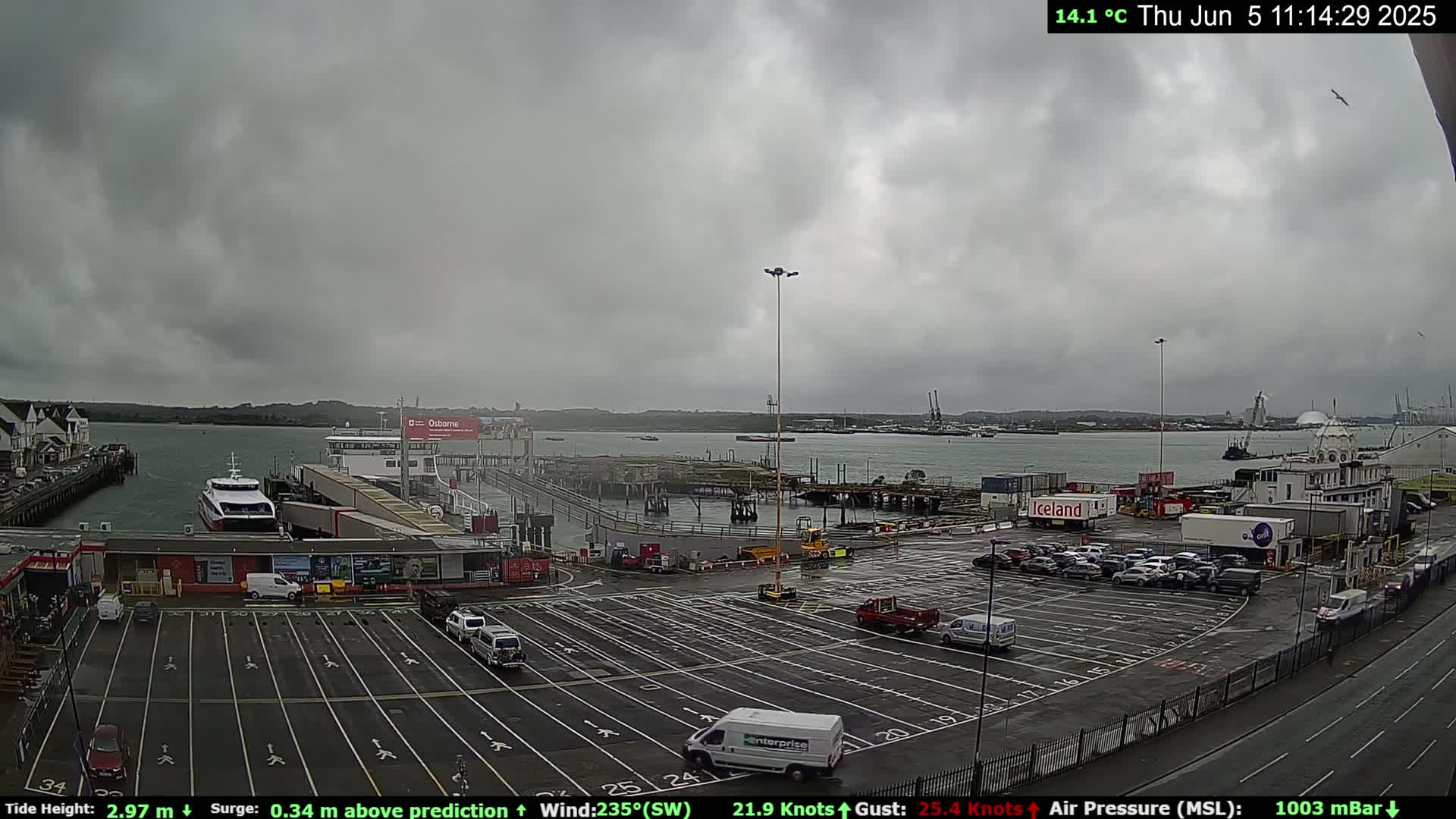 A ferry terminal with several cars parked in the lot, a ferry docked, and overcast skies.