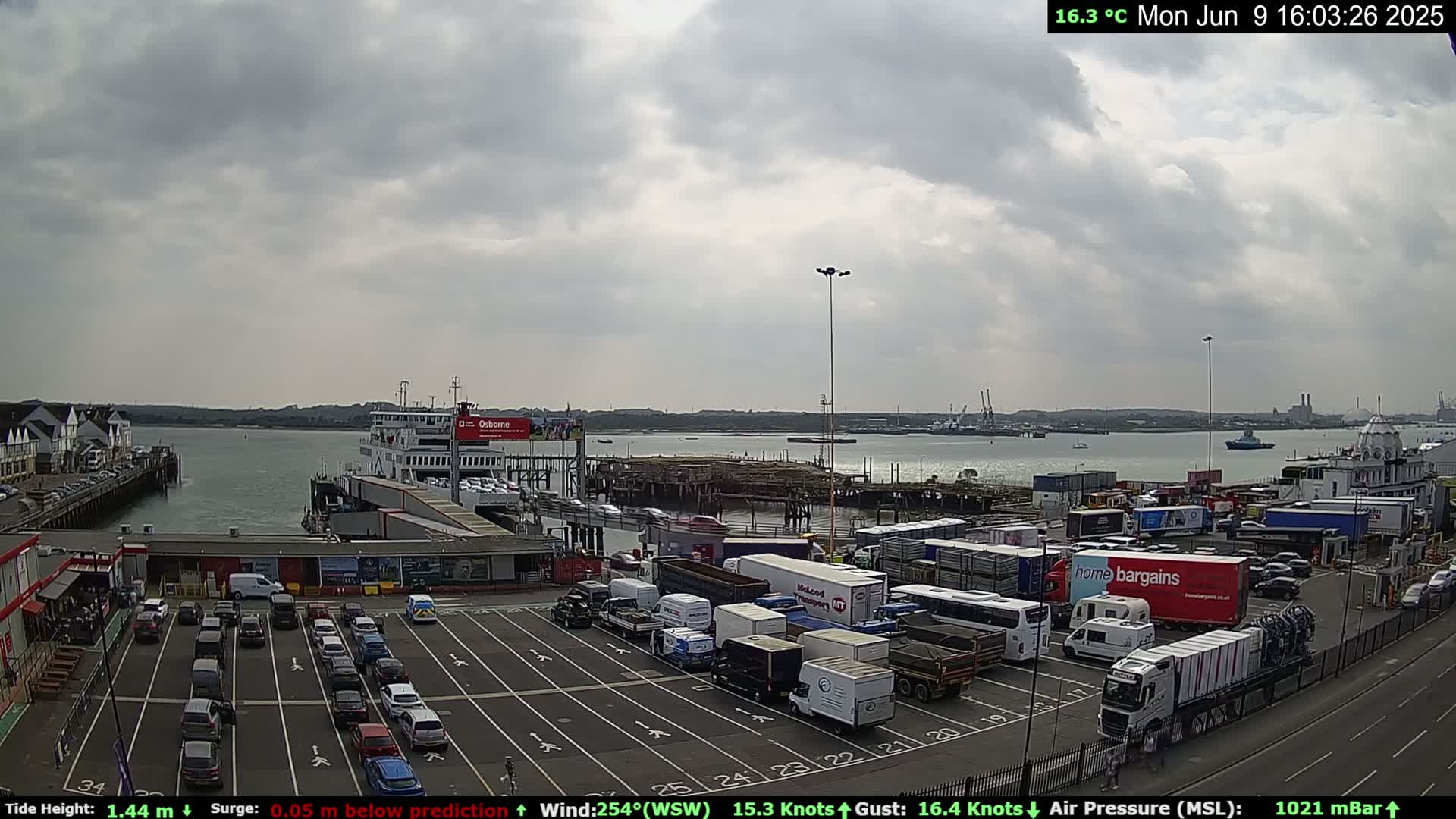 A ferry is docked at a terminal, with numerous vehicles parked in a lot and more vehicles parked along the waterfront under an overcast sky.