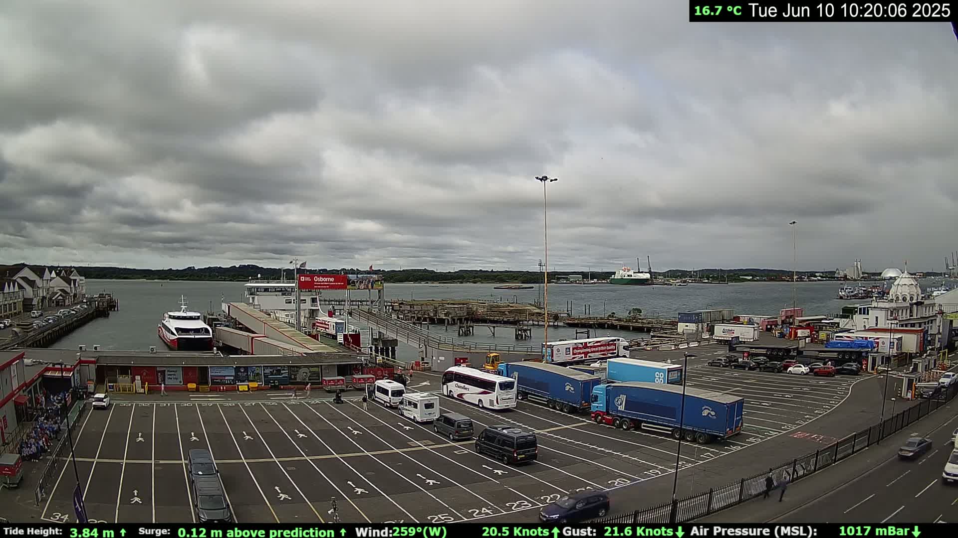 A ferry terminal with several vehicles parked, a ferry docked, and overcast skies.