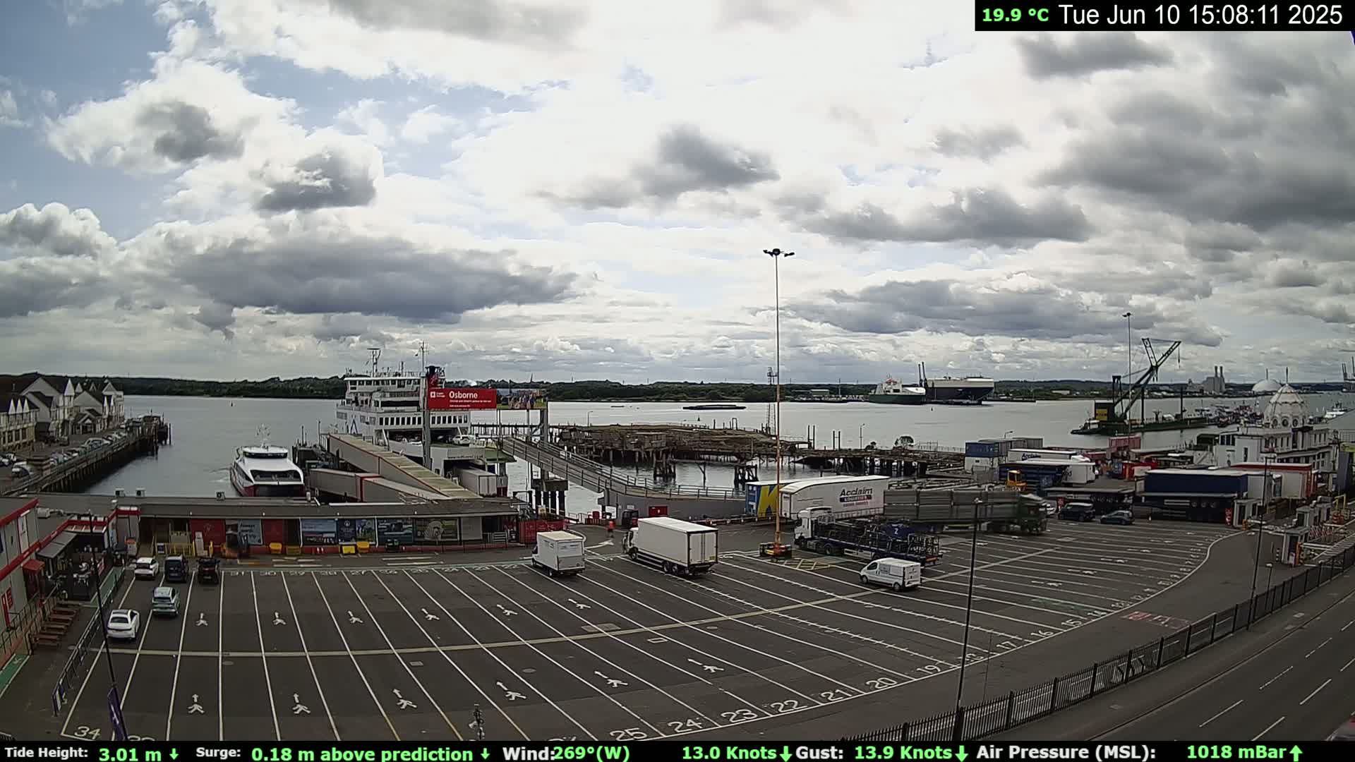 A ferry terminal with a large ferry docked, several smaller vessels in the water, and numerous trucks and cars parked in a lot under a partly cloudy sky.
