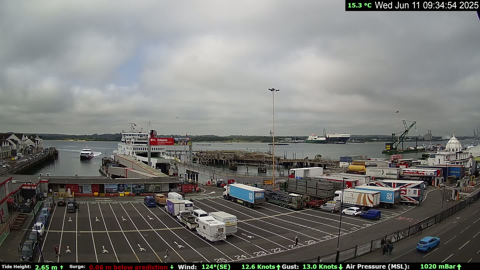 A ferry terminal with numerous trucks and cars in a parking lot, a ferry docked, and other vessels in the water under an overcast sky.