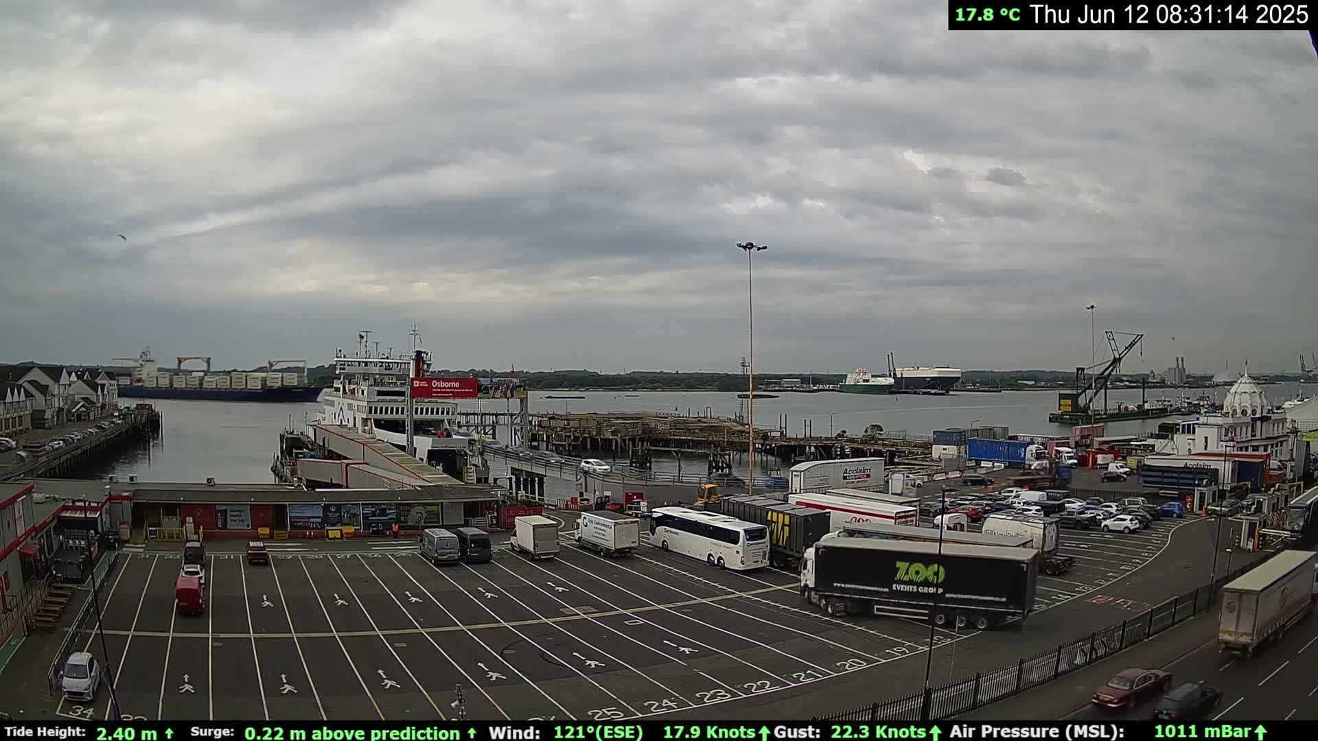 A ferry is docked at a port under an overcast sky, with numerous vehicles parked in a lot nearby and other ships visible on the water.